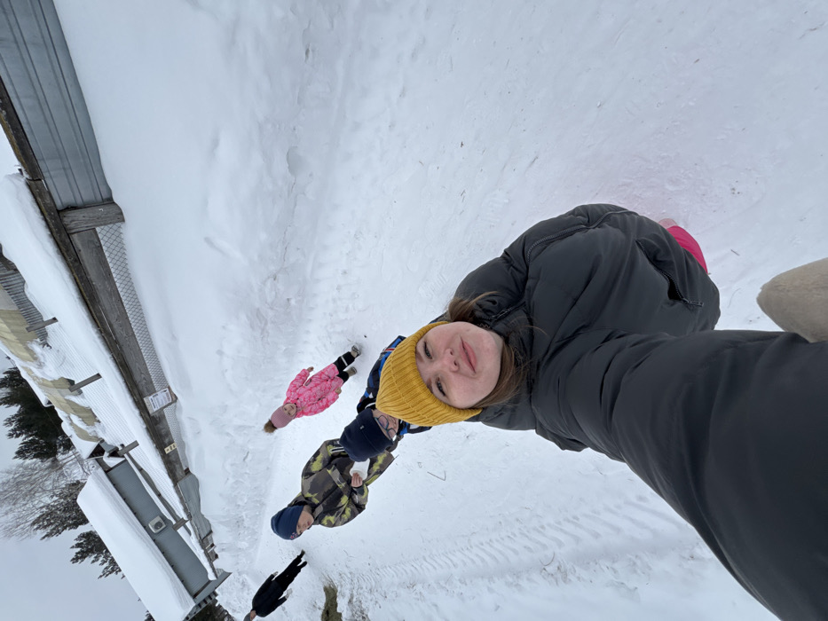 This image depicts a winter scene on a snow-covered path in Crokhovo, Russia, under an overcast sky. In the foreground, a young woman wearing a yellow knitted beanie and a black puffer jacket takes a selfie, smiling directly at the camera. Her right arm is extended, holding the camera, and a portion of a light-colored glove or sleeve is visible at the bottom.
Behind her, four individuals are visible walking along the snowy path. Immediately behind the woman, a boy wears a dark blue beanie and a camouflage-patterned winter jacket, holding a small white object, possibly a snowball or snow. To his right, a younger child, likely a girl, wears a bright pink puffer coat with a hood and black boots. Further behind the first boy, another child or individual with a dark beanie and a blue jacket is partially visible. Further in the background on the left, a person in a black coat and dark pants walks away from the camera.
The path is entirely covered in white snow, with discernible tire tracks on the left side. To the right of the path, a high bank of snow parallels the walkway. Beyond this snow bank, a fence line composed of wooden posts, horizontal wooden planks, and wire mesh is visible. Behind the fence, several small, utilitarian buildings with snow-covered roofs are present. In the distant background, snow-laden coniferous trees are visible beneath a uniform grey sky. A small pile of dried vegetation, possibly hay, is on the ground near the far-left individual.
