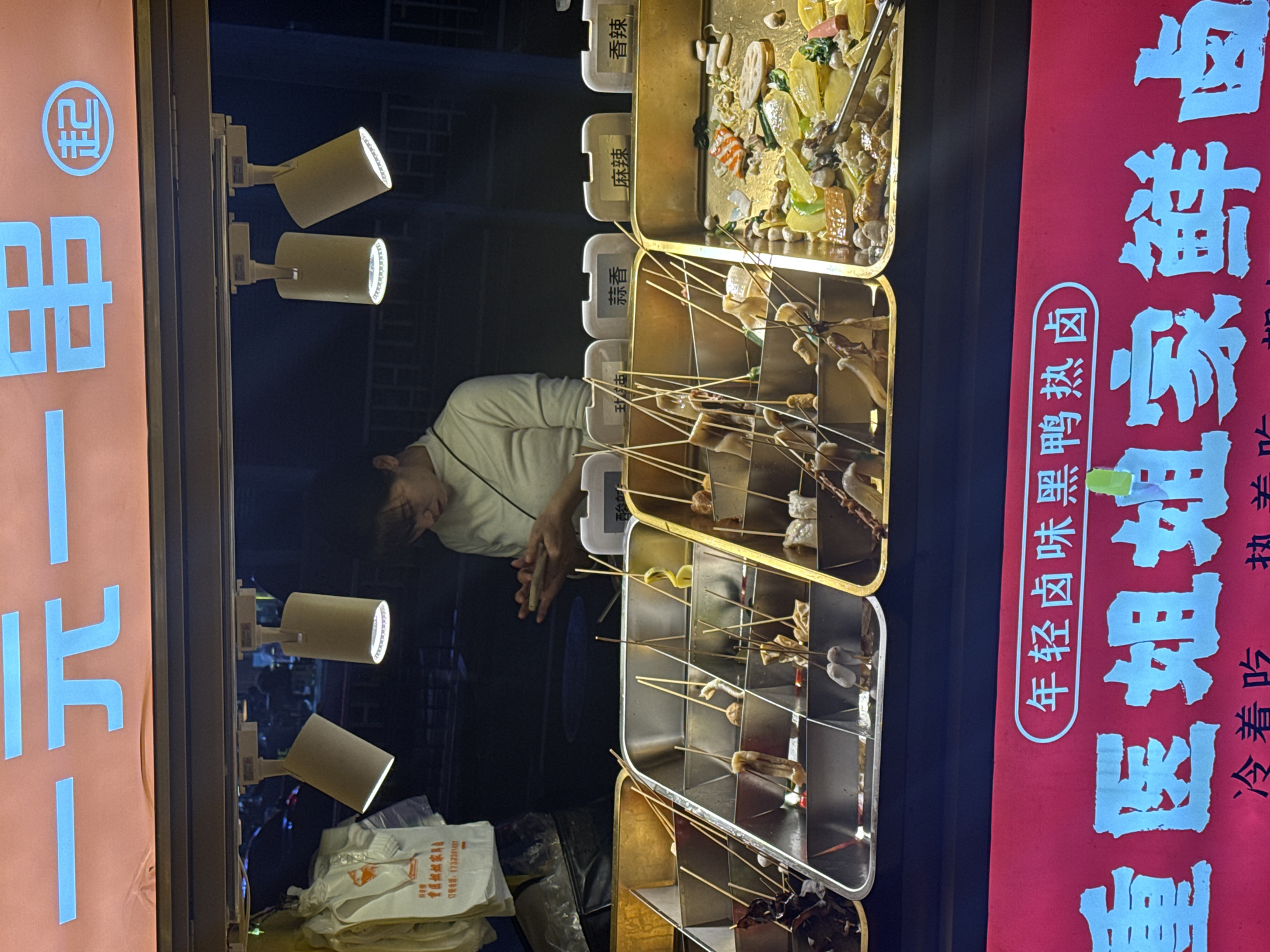 Here is a detailed description of the image:
**Setting and Environment:**
The image appears to be taken at a street food stall, presumably at night, in the Yuzhong District, China. The lighting is a mix of focused spotlights and ambient street lighting. The scene seems to focus on the food display and the person operating the stall.
**Subjects and People:**
The primary human subject is a person with short dark hair, presumably an adult, wearing a light-colored, collared shirt. They are standing behind a food display, with their hands clasped.
**Objects and Food:**
The food is presented on skewers in stainless steel trays. There are four distinct trays of food, labeled in Chinese as "Garlic Flavor," "Spicy," and "Fragrant Spicy." The topmost tray appears to contain vegetables and other foods cooked in sauce or broth.
**Signage and Writing:**
There is a red banner with white Chinese characters that translate to "Young People's Braised Duck." There is a smaller line of text that reads, "Sister's Spicy Duck" with instructions on serving temperature. Another sign advertises phone number for placing orders.
**Action:**
The person behind the stall is likely waiting for or attending to customers. The food display is actively lit and ready for selection.
**Overall Impression:**
The scene is a snapshot of street food culture, focusing on ready-to-eat food items presented in a clean and orderly display.