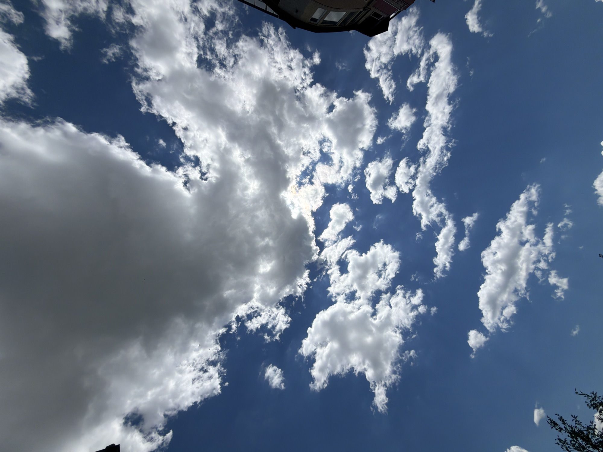 The image depicts a bright blue sky filled with various types of clouds, ranging from wispy cirrus to larger cumulus formations. The sun is partially obscured by a large, dark cloud in the upper portion of the frame, casting shadows and creating a dramatic contrast. Several smaller, fluffy white clouds are scattered across the sky. In the far right of the image, the corner of a multi-story building with windows is visible. In the lower left, the dark silhouettes of tree branches can be seen. The perspective is looking upwards towards the sky. This scene is consistent with a view from an urban environment, potentially in Burgas, Bulgaria, given the context. There are no visible people or specific identifiable landmarks that would pinpoint the exact location within the city.