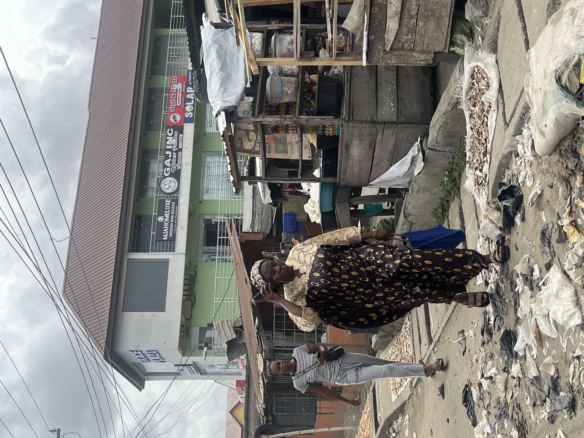 The image shows a street scene in Lekki, Nigeria. A woman in a brown and gold patterned dress stands on a sidewalk, holding a phone to her ear. Another person in a striped shirt and gray pants walks beside her. The sidewalk and street are littered with trash. In the background, there's a multi-story building with businesses, including "MJAHOMELUXE" and "GAJINC." A wooden structure stands next to the building, possibly a roadside stall. Overhead power lines are visible against a cloudy sky.