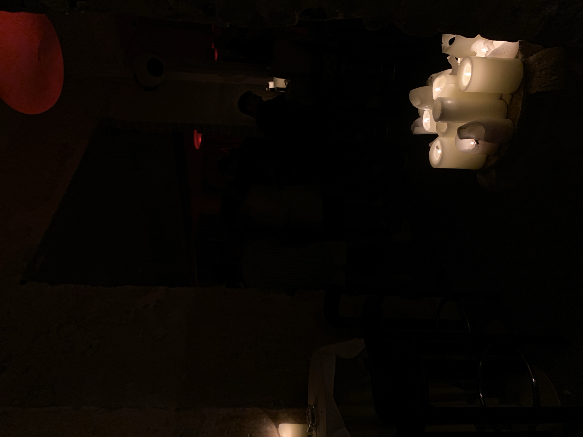 A dimly lit interior scene, possibly a bar or lounge, is captured in this photograph. The foreground is dominated by a cluster of lit white pillar candles of varying heights, casting a warm glow on their immediate surroundings. They are arranged on a rough, light-colored surface, likely a stone or wooden base.
To the left, a table is draped with a white tablecloth, and a single lit candle can be seen on its surface, along with what appears to be a lighter and cigarette butts. A dark stool with metal accents is partially visible in the lower left corner.
The midground and background are predominantly dark, with the silhouettes of people visible in the distance. Red lighting is present in some areas of the background, suggesting accent lighting or a red glow from further within the establishment. A circular red object, possibly a light fixture or decoration, is visible in the upper right portion of the image. A security camera is mounted on a wall in the mid-right background.
The overall ambiance is intimate and atmospheric, with the soft light of the candles creating a sense of warmth and mystery. There is no visible indication of the location being in Tel-Aviv, Israel.