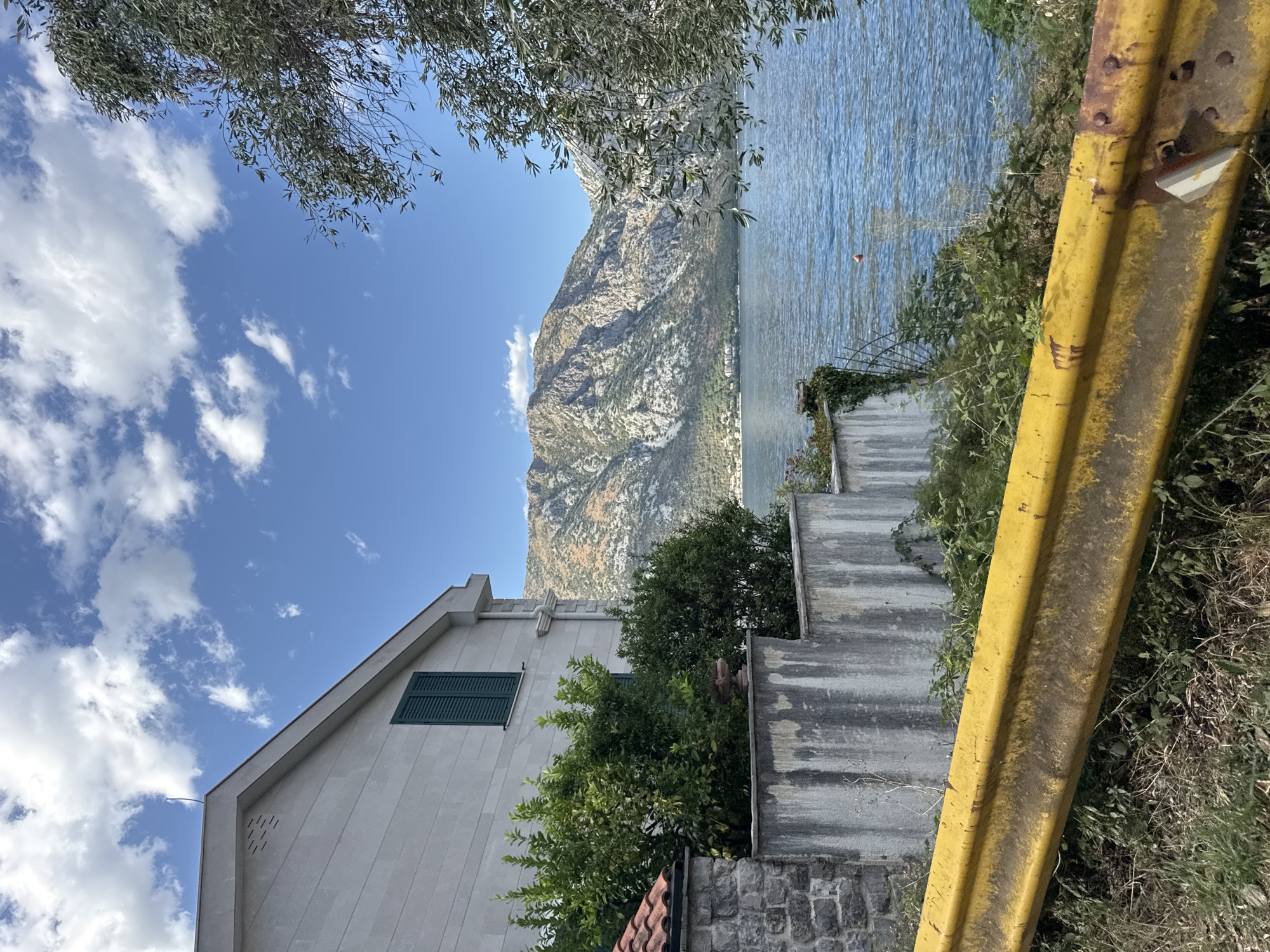 The image depicts a seaside view in the town of Prčanj, Montenegro. In the foreground, a weathered yellow guardrail runs diagonally across the frame, bordered by dry grass and weeds. To the left, a light-colored building with a tiled roof and a green shuttered window stands next to a stone wall. Beyond the building and wall, dense green foliage grows, obscuring the base of a steep, rocky mountain that dominates the background. The mountain slopes down to meet the calm, blue water of a bay. The water reflects the bright blue sky, which is dotted with white, fluffy clouds. A small, orange buoy is visible floating on the water in the middle distance. The scene is likely captured on a sunny day, offering a picturesque view of the Adriatic coastline.