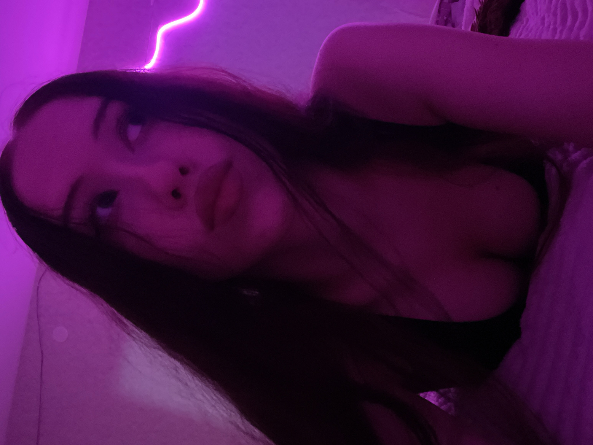 The image is a close-up portrait of a young woman lying down, illuminated by purple neon lights. Her face is turned to the side, with her gaze directed upwards and away from the camera. Her long, dark hair is spread out around her. She is wearing a dark top that reveals her cleavage. The background is dimly lit, with a prominent neon light strip on the right side of the frame. The setting appears to be a bedroom, with a textured surface, possibly a bed, visible in the foreground. The overall mood is intimate and atmospheric, due to the low lighting and the color of the neon lights. There is no information in the image that can identify the location as Votkinsk, Russia.