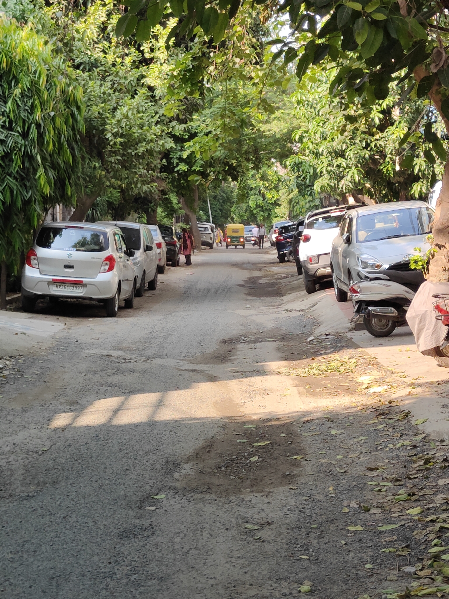 The image depicts a narrow, paved road lined with parked cars and lush green trees, suggesting a residential or suburban area. The road surface appears worn and uneven, with patches of dirt and scattered fallen leaves. On the left side of the road, a silver hatchback with license plate "HR26CC3997" is parked, followed by other vehicles. On the right side, a silver SUV and a scooter are parked. In the distance, a yellow auto-rickshaw and a few pedestrians are visible, indicating movement and activity. The scene is bathed in sunlight, casting shadows from the trees onto the road, creating a dappled effect. The overall impression is of a typical street scene in Gurgaon, India, characterized by a blend of urban infrastructure and abundant vegetation.