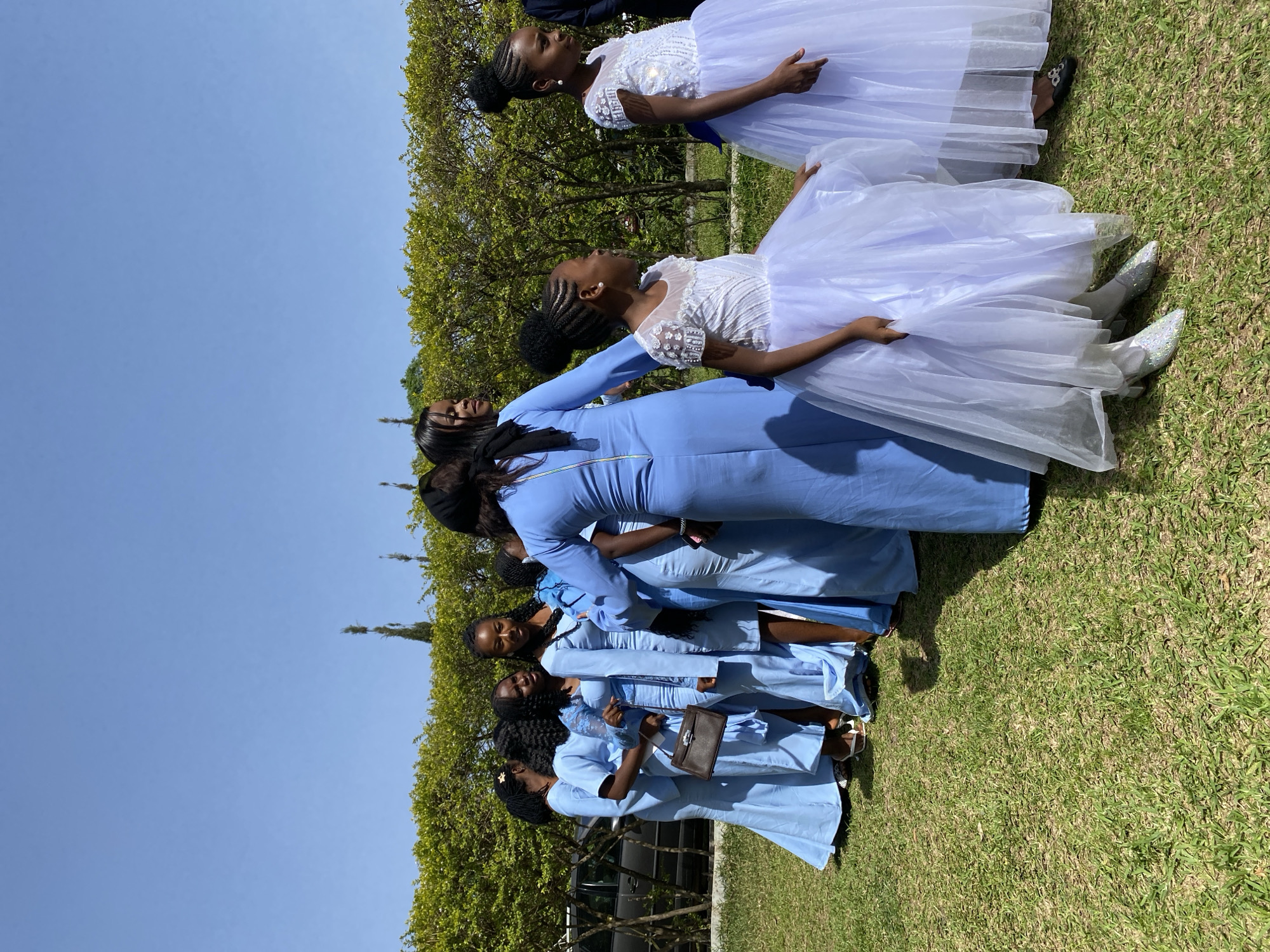 A group of women and young girls are standing on a grassy field under a clear blue sky, near what appears to be a hedge. The women are wearing light blue dresses, while the girls are in white dresses with tulle skirts. One of the women is carrying a dark handbag. Based on the location provided, this scene is likely in or near Bukuru, Nigeria.