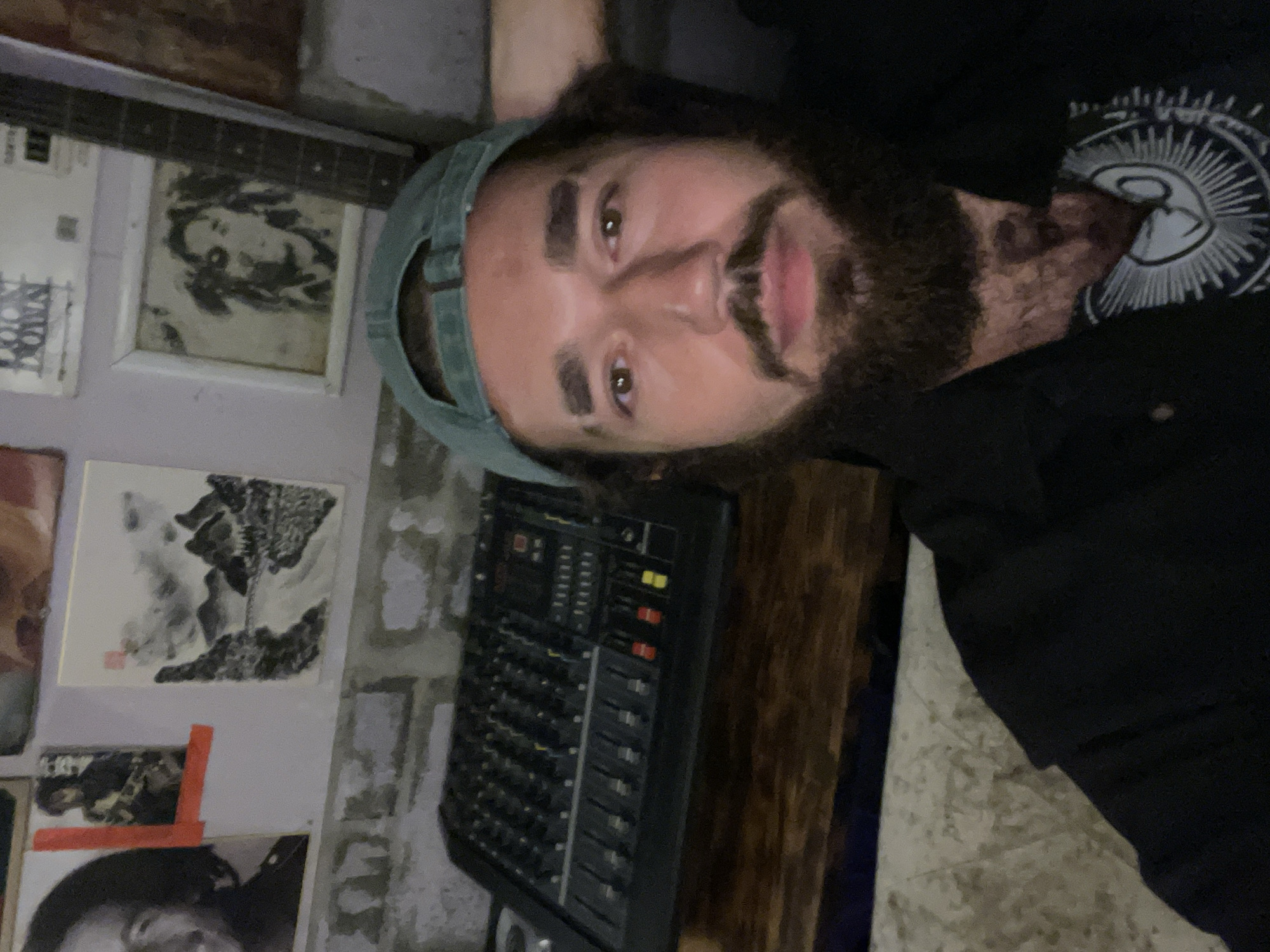 A man with a beard and mustache, wearing a green baseball cap and a dark shirt, is taking a selfie in a room. Behind him, there is a mixing board on a wooden desk. The walls are decorated with various framed pictures, including a painting of mountains, a picture of Bob Marley, and a photo of a person playing a guitar. A portion of a guitar neck is visible on the right side of the image. The room appears to be a personal studio or living space, possibly in Chalong, Thailand.