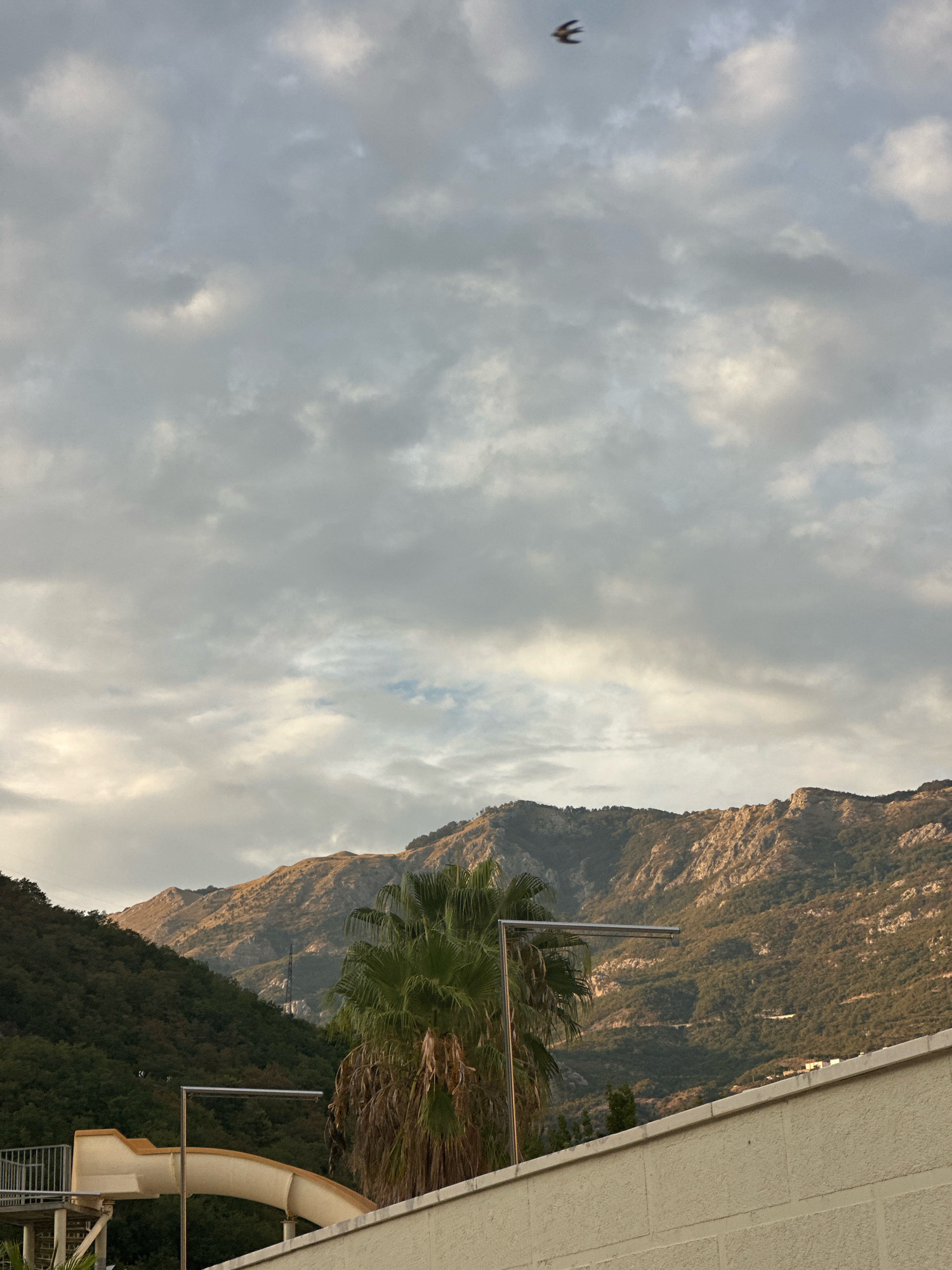 The image depicts a cloudy sky over a mountainous landscape in Boreti, Montenegro. In the foreground, a portion of a building with a cream-colored textured wall is visible, along with the top of a yellow water slide structure and a metal railing. A palm tree stands to the right of the slide. The mid-ground is dominated by lush green hillsides and rugged, sun-drenched mountains in the distance, with some sparse vegetation on their slopes. A single bird is captured in flight in the upper portion of the sky. The overall scene suggests an outdoor recreational area situated near a mountainous region under overcast conditions.