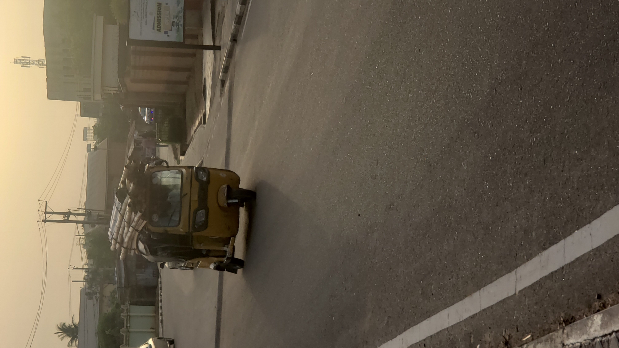 A yellow auto-rickshaw, heavily laden with goods under a tarpaulin cover, is positioned on a paved road with a white line marking the edge. The setting appears to be an urban area in Unknown, Unknown, suggested by the presence of buildings, power lines, and what appears to be an advertising sign on a structure to the left. The sky is slightly hazy, possibly due to early morning light or pollution. No people are clearly visible, although one may be driving the auto-rickshaw.