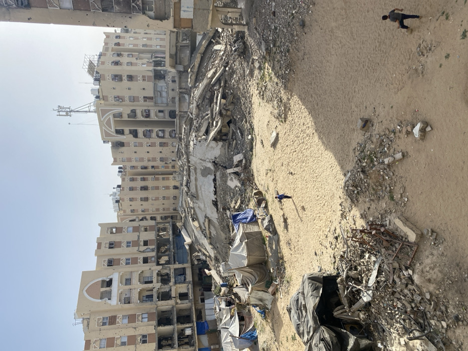 This image displays a scene of widespread urban destruction in Khan Yunis, Palestinian Territory. The central area features a massive, irregularly shaped pile of pulverized concrete and structural debris, indicative of a collapsed multi-story building. Surrounding this central rubble are areas of sandy ground, interspersed with smaller accumulations of debris, including concrete fragments and twisted rebar.
To the left, approximately eight makeshift shelters constructed from various tarpaulins and fabric are clustered together. Two individuals are present on the sandy ground: one person wearing a blue shirt walks from left to right in the mid-ground, casting a distinct shadow, and another person dressed in dark clothing is partially visible in the bottom right corner, standing near sparse debris.
In the background, several multi-story, beige-colored apartment buildings stand heavily damaged. These structures exhibit numerous impact holes, shattered windows, partially collapsed walls, missing balcony sections, and visible charring. Architectural features, including arched entryways and decorative elements, are present on some of the compromised buildings. A communications antenna tower is visible on the roof of one building in the center-right background. The sky is clear and light blue, and the scene is illuminated by bright daylight, casting sharp shadows.