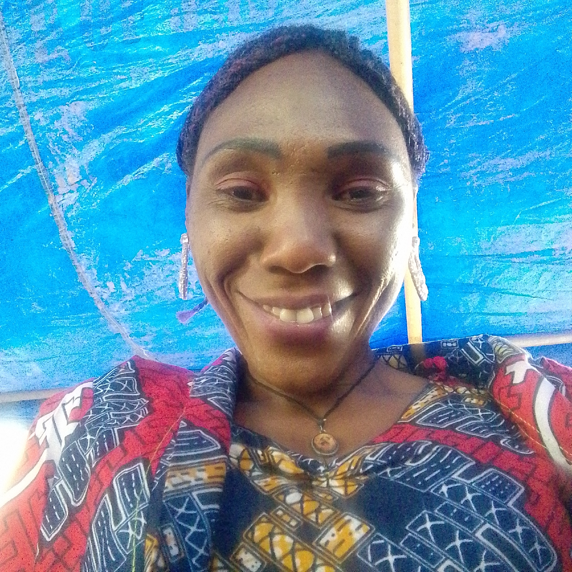 A close-up, medium shot depicts a Black woman smiling at the camera. The woman is adorned with earrings and a necklace featuring a circular pendant. She is wearing a multi-colored patterned shirt, predominantly red, navy, and yellow.
The backdrop consists of a blue surface, likely a tarp, with white streaks and some poles visible. The location is not evident and therefore described as Unknown, Unknown.