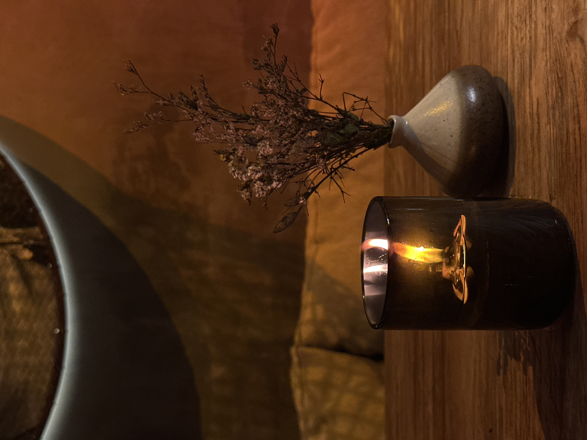 A dimly lit interior, possibly a bathroom in Pererenan, Indonesia, features a contemporary bathtub with a curved edge partially visible on the left. A lit candle in a dark glass container and a small ceramic vase holding dried flowers are positioned on a wooden surface, likely a shelf or counter. The background shows warm, earthy tones, contributing to the intimate ambiance.