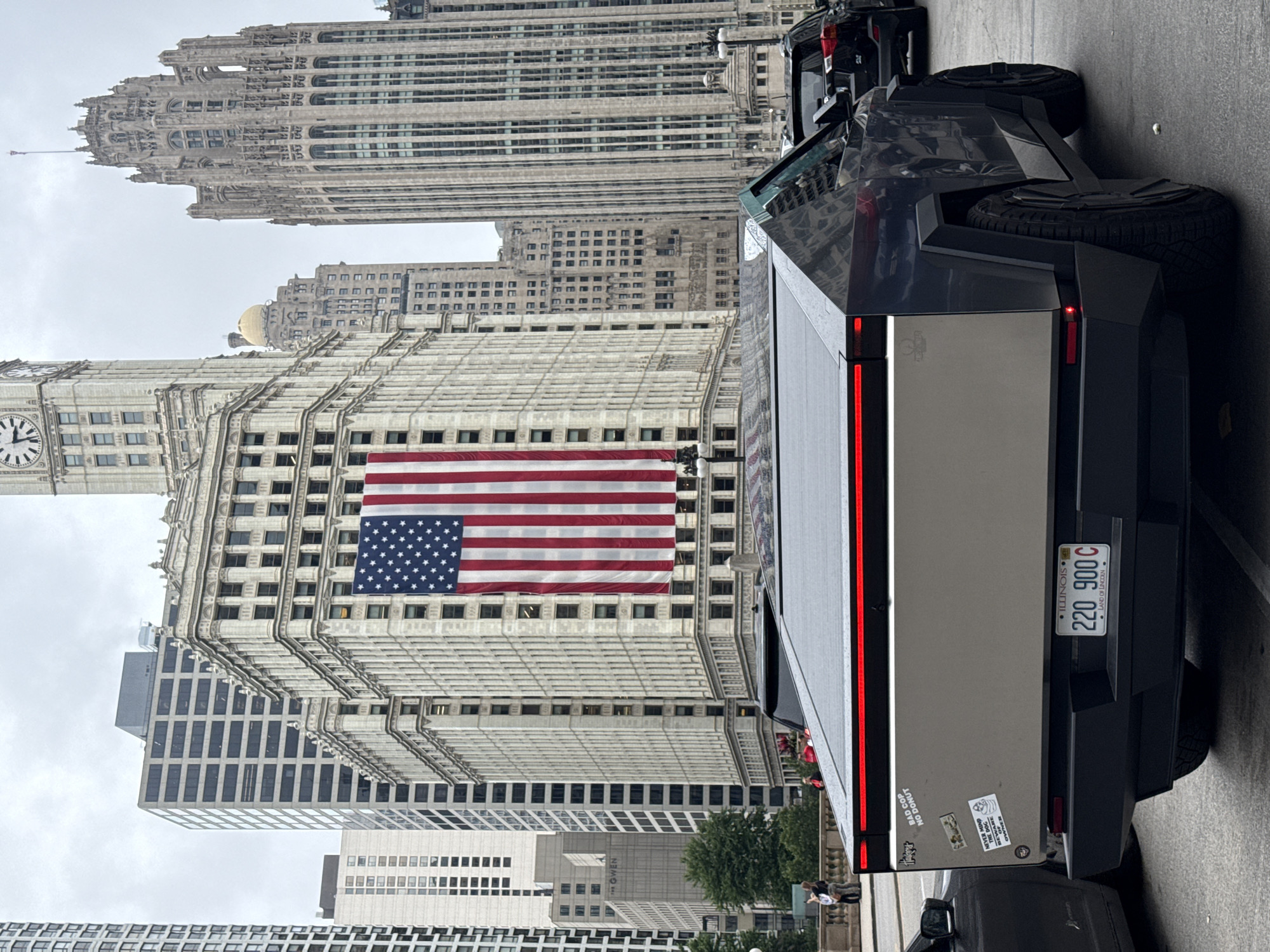 A Tesla Cybertruck, with a license plate that reads "220 900 C LAND OF LINCOLN", is parked on a street in Chicago. The vehicle's rear is visible, featuring a matte silver panel with several stickers. One sticker says "BAD COP NO DONUT", and another reads "NEVER MIND THE DOG, BEWARE OF THE OWNER". The truck is parked in front of several large buildings, including the Chicago Board of Trade Building, which is adorned with a large American flag. Other skyscrapers are visible in the background, under a cloudy sky. People are seen walking on a sidewalk to the left of the truck.