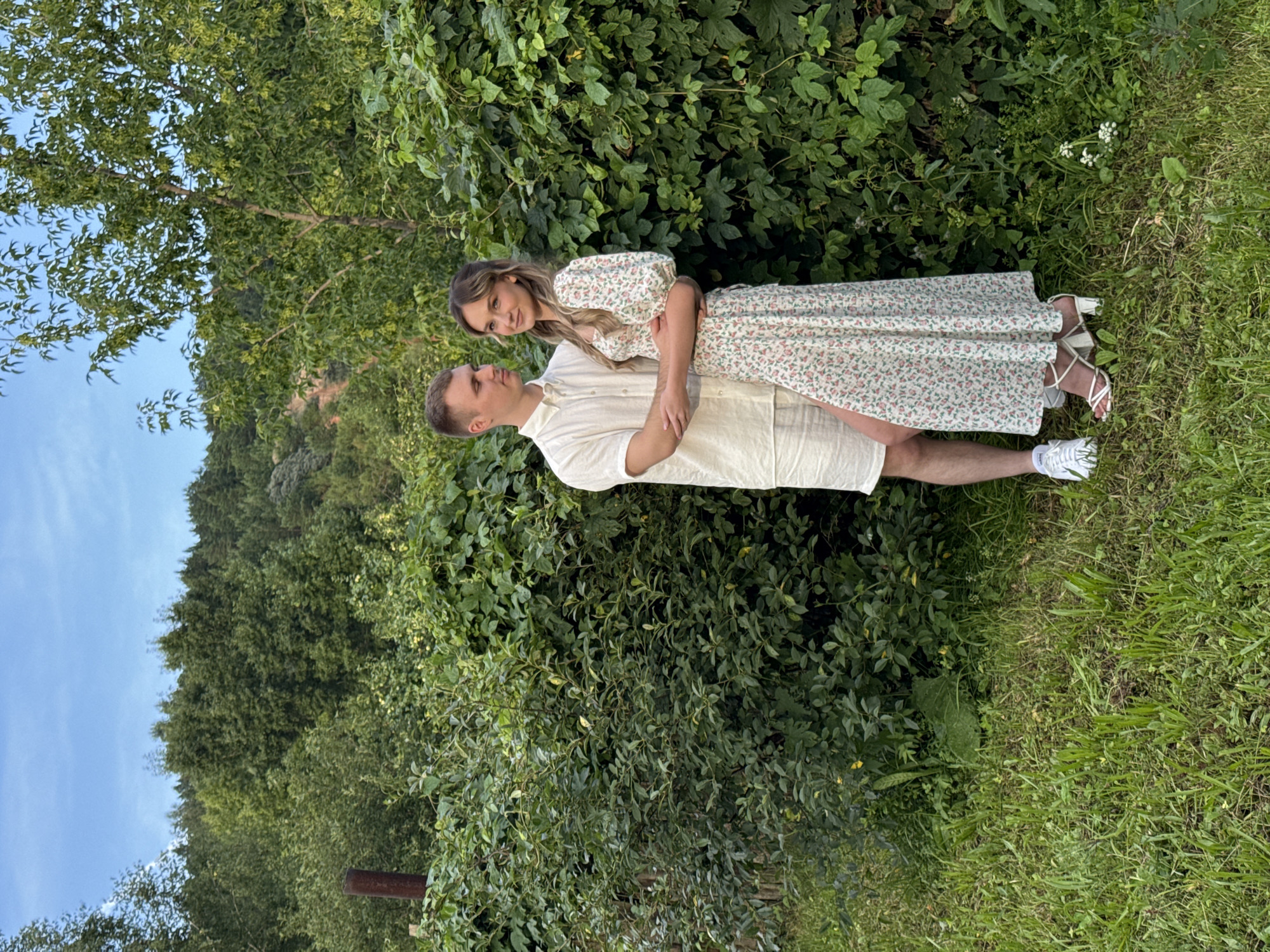 An outdoor, medium shot captures a young Caucasian couple embracing near a dense, green hedge. The man, on the left, wears a short-sleeved, cream-colored button-up shirt and matching shorts. He has short, brown hair and is looking towards the woman. The woman, on the right, sports a long, cream-colored dress adorned with a floral pattern of pink and green. Her hair is long and blonde. She is looking at the camera. Her arms are around the man's neck and waist, while his arm is around her waist. Both are wearing white shoes. The background features a lush, green hedge, overgrown grass, and trees under a partly cloudy sky. The scene appears to be a rural or suburban setting, potentially in or near the city of Olkhovka, Russia, given the presence of abundant greenery and the overall rustic aesthetic.
