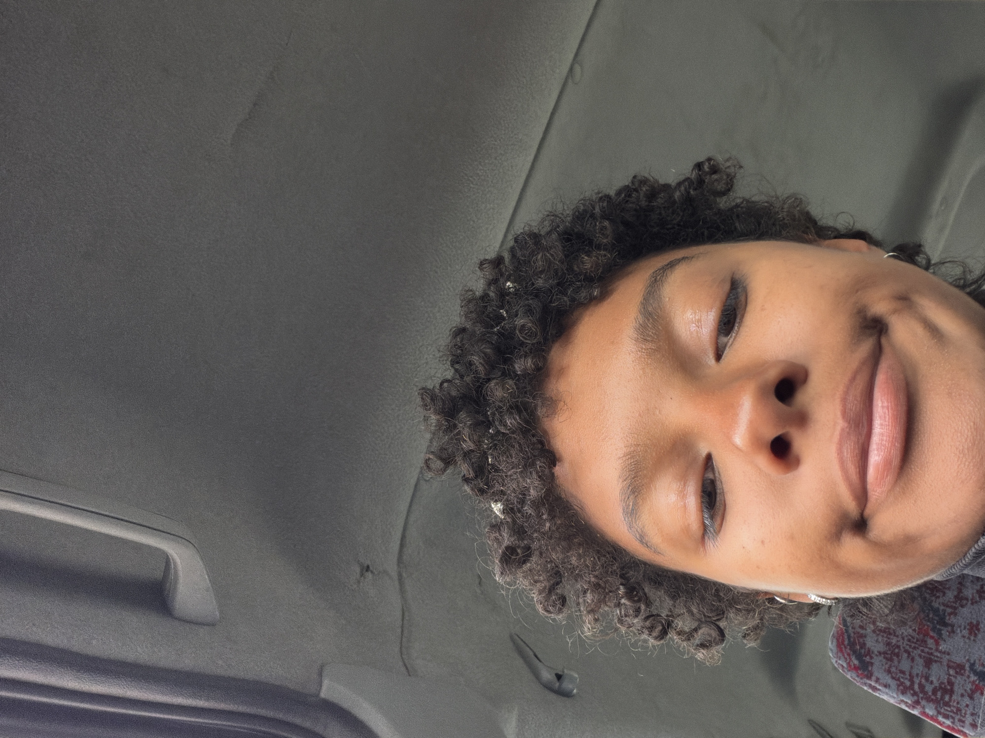 The image shows a close-up, angled perspective of a person's face, appearing to be taken from a low angle, possibly inside a vehicle. The person has curly hair and is partially framed against a gray, textured surface. A handle is visible in the lower left corner, suggesting an interior car fixture. The person seems to be looking down towards the camera, exhibiting a subtle smile. The environment appears to be the inside of a vehicle, not a location in the city of Pals, Spain.