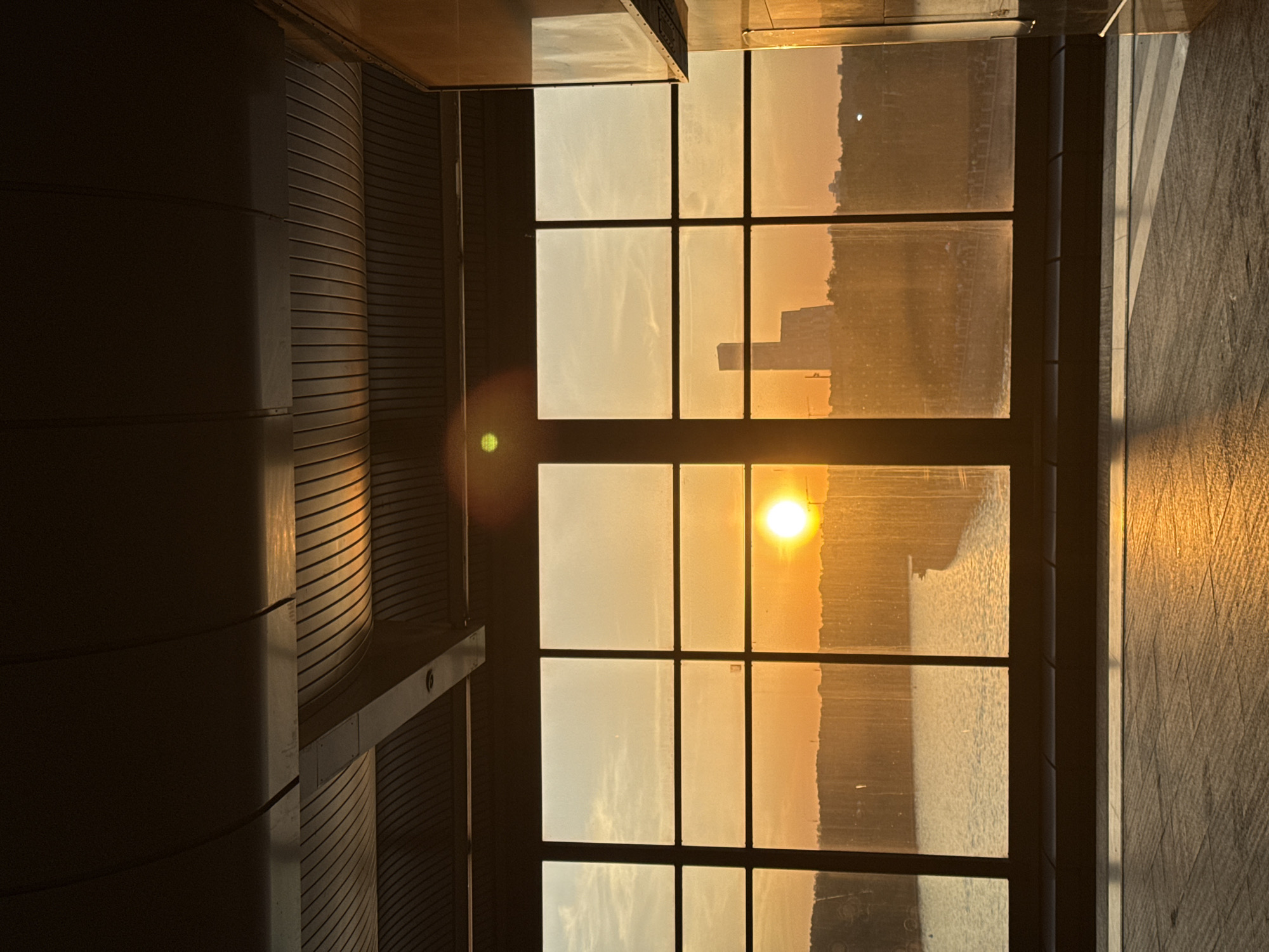 The image depicts a sunset view through a window with a grid pattern. The sun is visible through one of the panes, casting a bright glare. Outside the window, a body of water and the skyline of a city are visible. The city likely includes high-rise buildings. To the left of the window is a section of the building's interior, possibly part of a curved wall or architectural feature. The window frame is black, and the surrounding wall appears to be made of stone. The location is in Moscow, Russia.