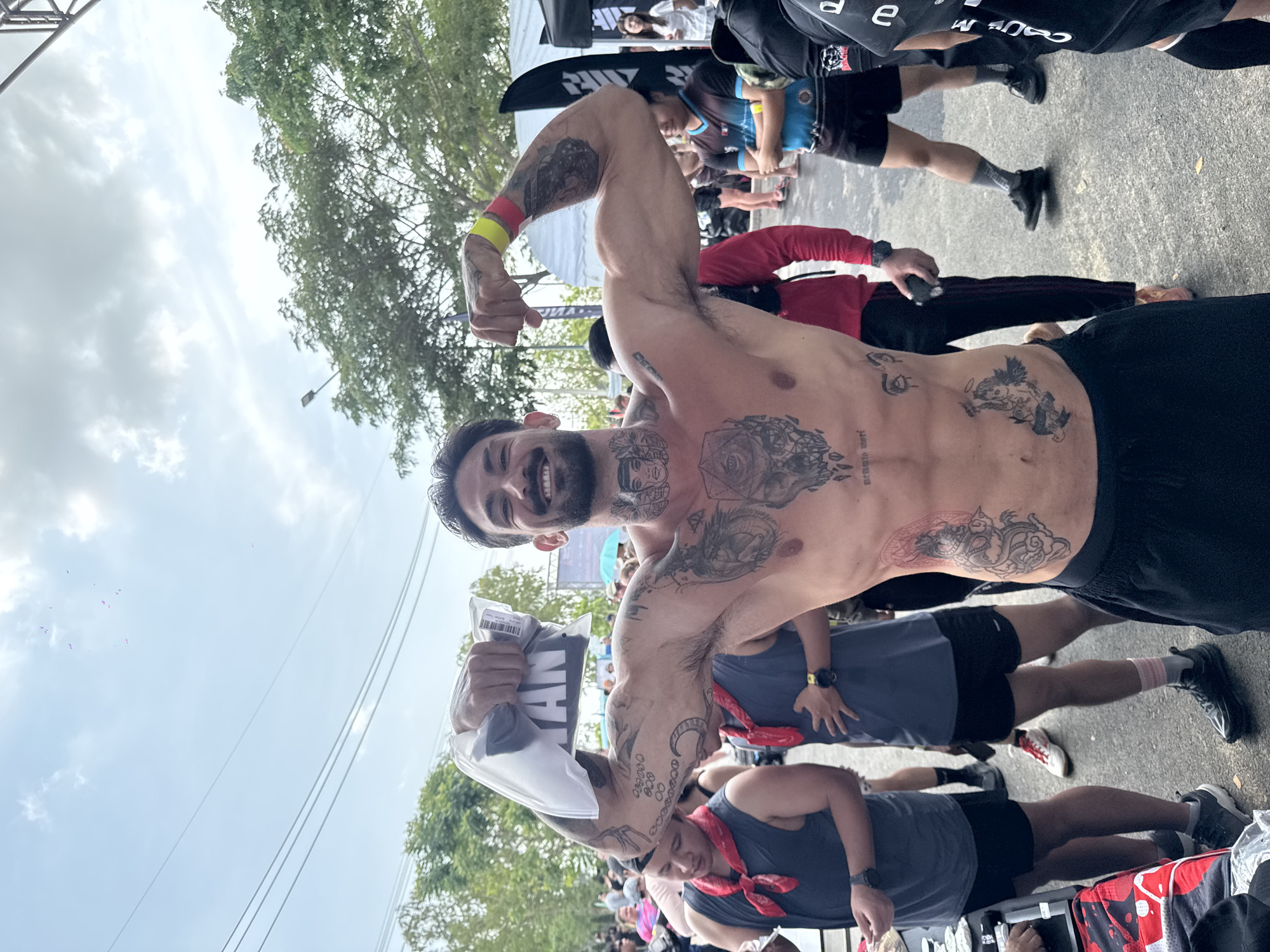 The image shows a shirtless man with multiple tattoos flexing his arms, likely at a race or event. He is holding a packaged item with the word "MAN" visible on it. The setting is likely at the finish line of the race in the city of บ้านสำนักตะแบก, Thailand, due to the presence of other participants and event-related structures nearby. The man is surrounded by other runners, some wearing tank tops with red bandanas. The weather appears overcast.