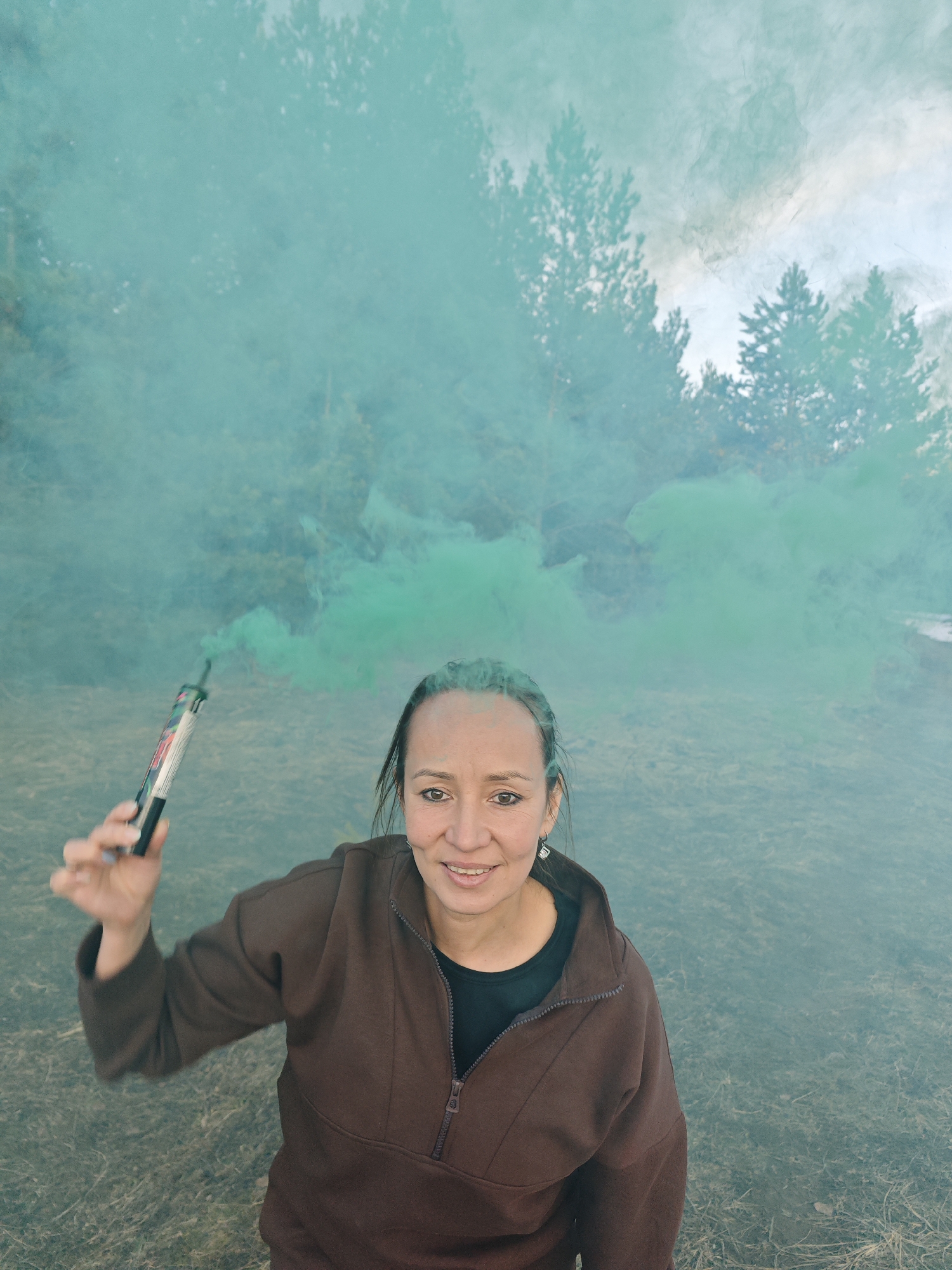 A medium-shot portrait captures a woman outdoors in Dubovaya, Russia, holding an activated green smoke grenade. The woman, with a slight smile, looks directly at the camera. Her face shows some green residue, likely from the smoke. She is wearing a brown, long-sleeved, collared top with a partially unzipped front, revealing a black undershirt. Small earrings are visible.
The smoke grenade, held in her right hand, emits a dense plume of vibrant green smoke that billows around her and into the background, obscuring parts of the scenery. The background features a forest of pine trees, partially veiled by the smoke, under what appears to be an overcast or hazy sky. The ground is covered with dry, sparse grass. The overall impression is a daytime scene, possibly during late afternoon, with a playful or experimental tone due to the use of the smoke flare. Text and imagery are visible on the smoke grenade's label, but they are too small to be deciphered.