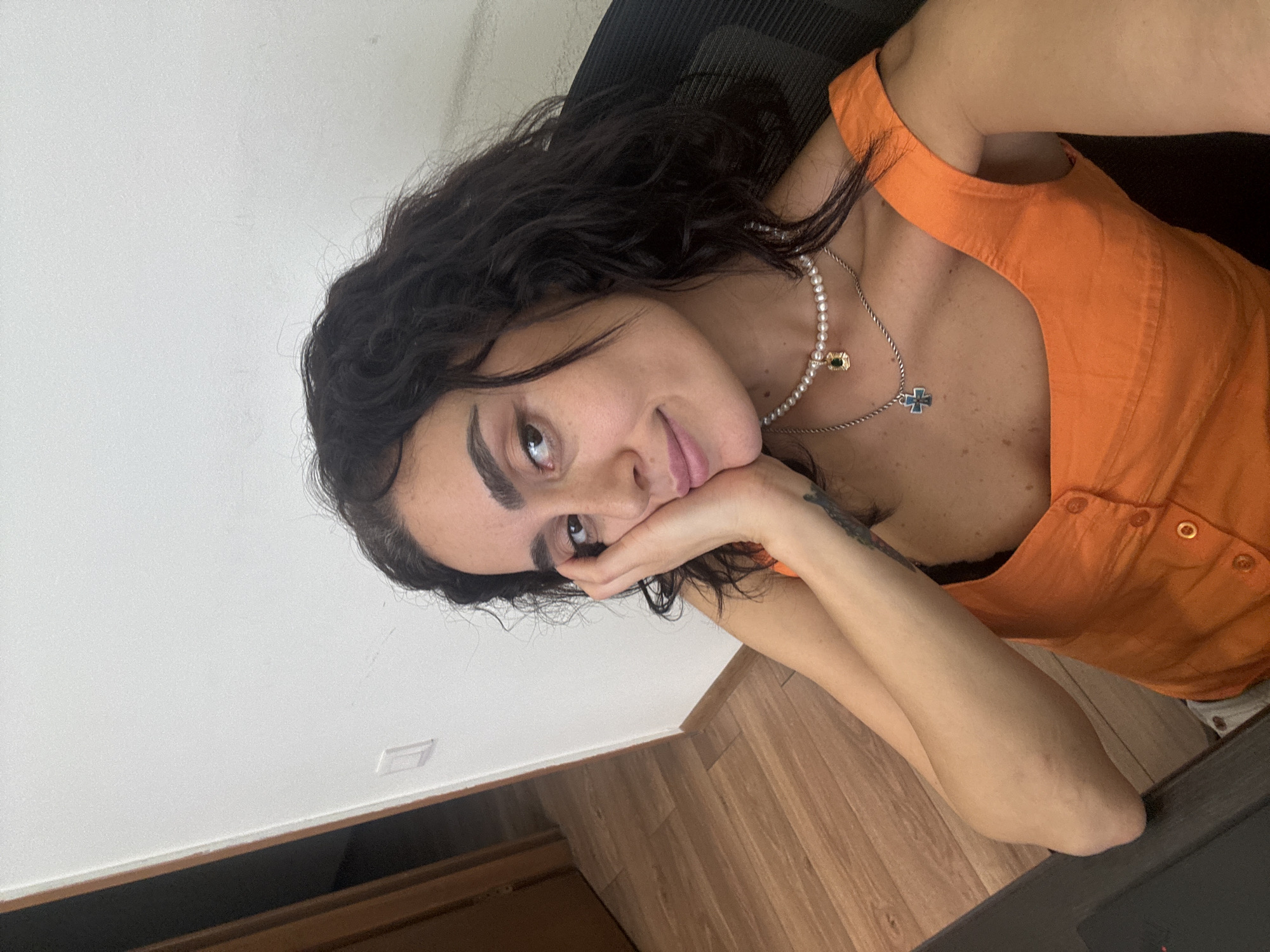 The image shows a close-up of a woman with dark, curly hair and fair skin, resting her head on her hand. She is wearing an orange top and two necklaces, one with pearls and a green pendant, and another with a blue cross pendant. She appears to be indoors, in what looks like an office or a room with wooden floors and a white wall. The background shows a doorway and a part of a chair. There are no visible indicators in the image to suggest the location is Tbilisi, Georgia.