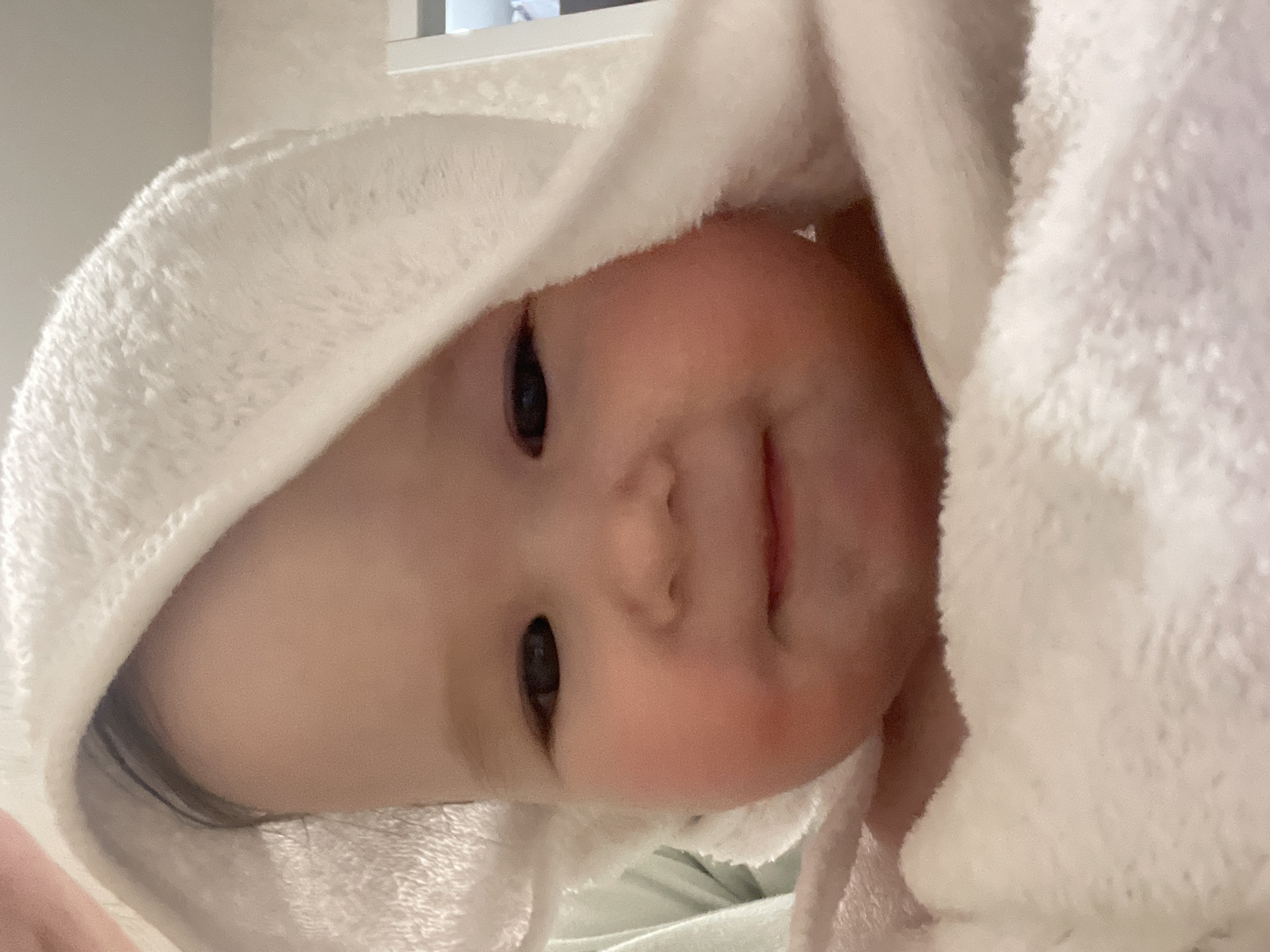 A close-up, slightly angled portrait shows a baby swaddled in a white, fluffy hooded towel. The baby's face, with rosy cheeks, dark eyes, and a slight smile, is the focal point. Some of the baby's dark hair is visible at the edges of the towel. The background is out of focus and appears to be a room interior with a window frame visible in the upper part of the image. It is not possible to determine the location in the city of Chaikovsky, Russia from the provided image.
