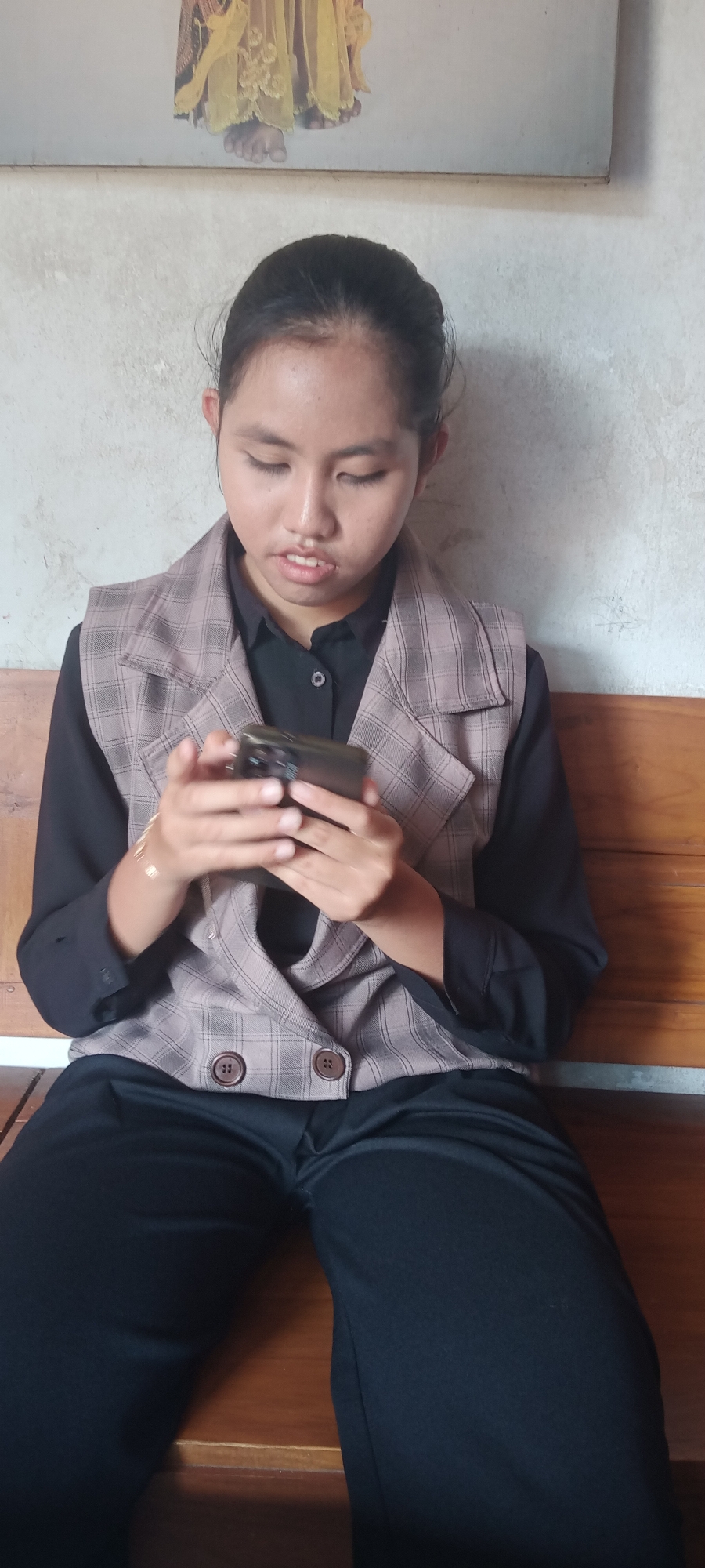 In Singgahan, Indonesia, a young woman sits on a wooden bench, engrossed in her smartphone. She is wearing a black collared shirt and a plaid vest with two prominent buttons. Her dark hair is pulled back. The background features a textured wall, possibly concrete, with a framed artwork depicting what appears to be a figure in yellow robes above her. The overall lighting suggests an indoor setting, likely during the daytime.