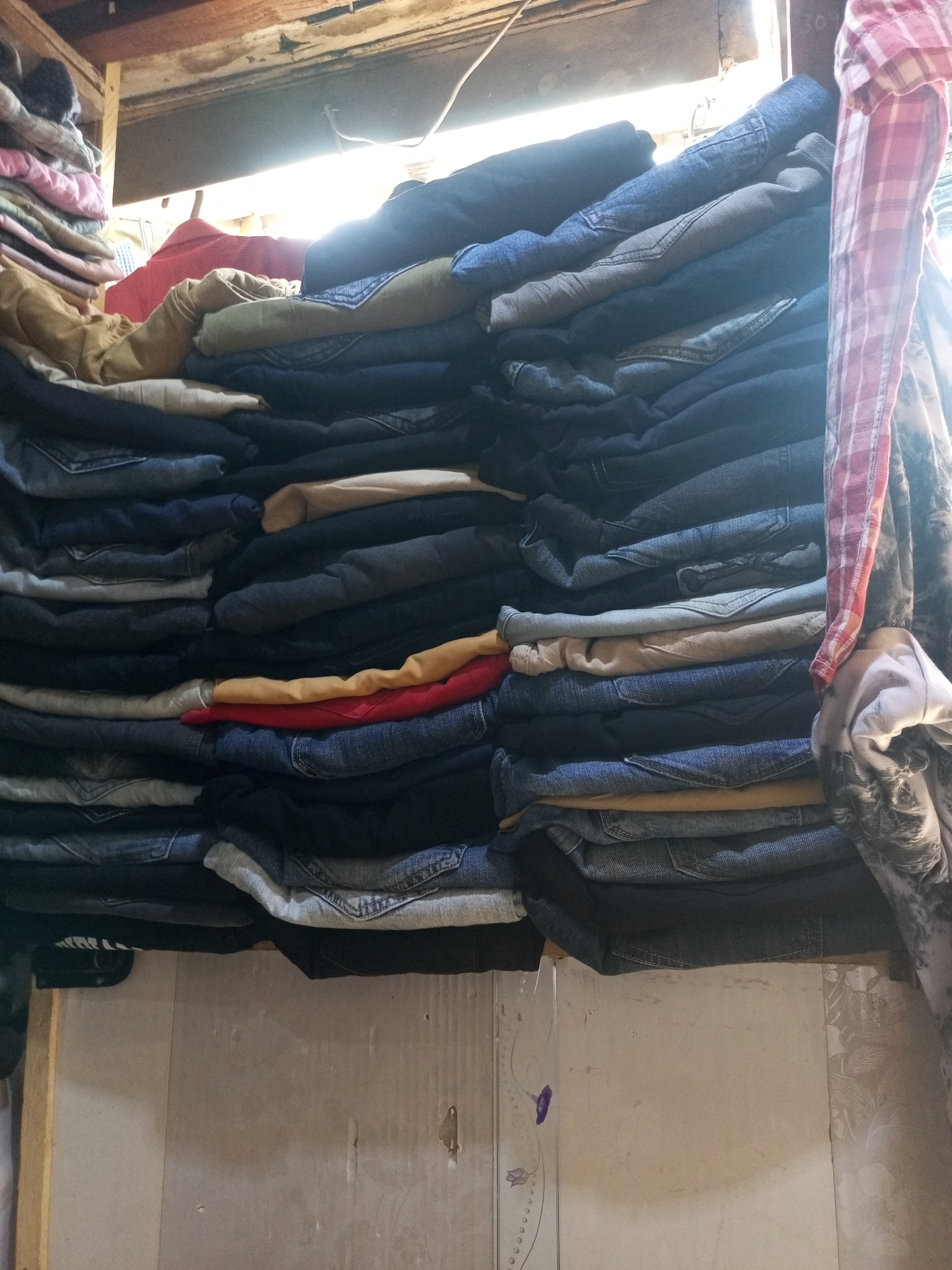 A stack of folded clothing, mostly jeans in varying shades of blue and gray, is piled on what appears to be a shelf or ledge. A few non-denim items, including pieces of red, yellow, and beige fabrics, are interspersed within the stack. To the right, a plaid shirt and a patterned cloth hang. The setting seems to be an indoor space with a wooden ceiling visible at the top of the frame. The location is Maiburiji, Nigeria.