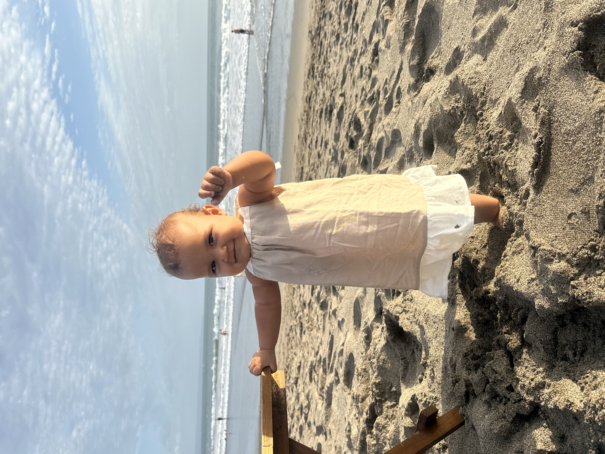A young child is standing on a sandy beach in North Kuta, Indonesia, with the ocean and sky in the background. The child, with curly brown hair, is wearing a light-colored dress with a white ruffled hem. They are facing the camera and smiling, with their right hand raised, holding a small amount of sand. To the left of the child, a wooden table or chair leg is partially visible. The sand on the beach shows footprints and is textured with shadows from the late afternoon sun. The ocean has gentle waves, and several people can be seen wading in the water in the distance. The sky is partly cloudy with a mix of blue sky and white clouds.