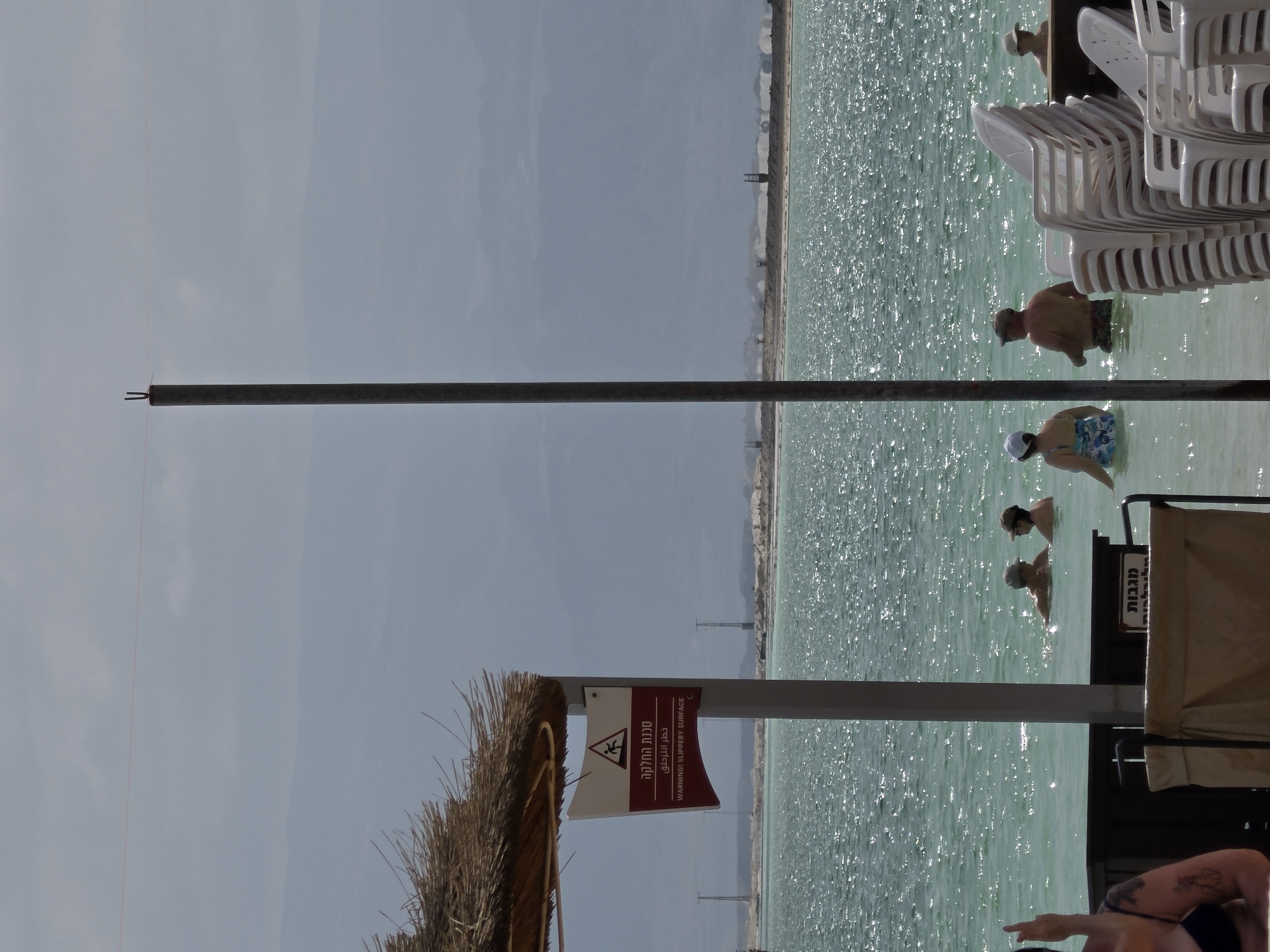 The scene depicts a waterside view in Tamar Regional Council, Israel. There is a body of water with several individuals wading in it. Stacks of white plastic chairs are positioned near the water's edge. A warning sign about a slippery surface is visible, and a thatched umbrella or shelter is in the foreground. A metal pole extends from the bottom to the top of the frame. The sky is overcast, and mountains can be seen in the distance.