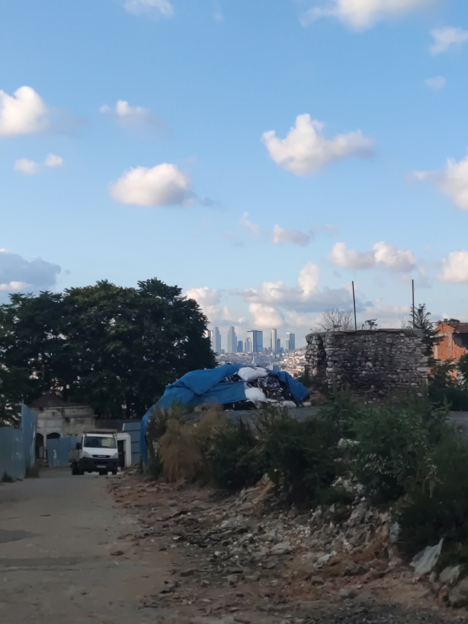 In this vertical, medium-shot image, a truck is driving down a dirt road in what appears to be a developing or industrial area of Istanbul, Turkey, with the city's skyline visible in the distance. The sky is a bright blue with scattered white clouds.
In the foreground, the road is unpaved and appears rough, with piles of debris and some vegetation on either side. To the left, a white truck with a flatbed is driving away from the camera. It is the only vehicle visible. Beyond the truck, a light-colored building with an arched entrance is partially visible, surrounded by blue construction barriers.
In the middle ground, a large pile of materials, covered by a bright blue tarp, is situated on the right side of the road. Behind this pile, a circular stone structure, possibly an old tower or well, stands with vegetation growing around its base. Further to the right, the corner of a red brick building with a damaged roof is visible.
Dominating the background is the Istanbul skyline, featuring numerous modern skyscrapers, suggesting the city's urban center is some distance away. To the left of the skyline, a large, dark green tree with dense foliage stands prominently. The overall impression is one of contrast between a raw, developing area and the sophisticated urban landscape beyond. No people are visible in the image.