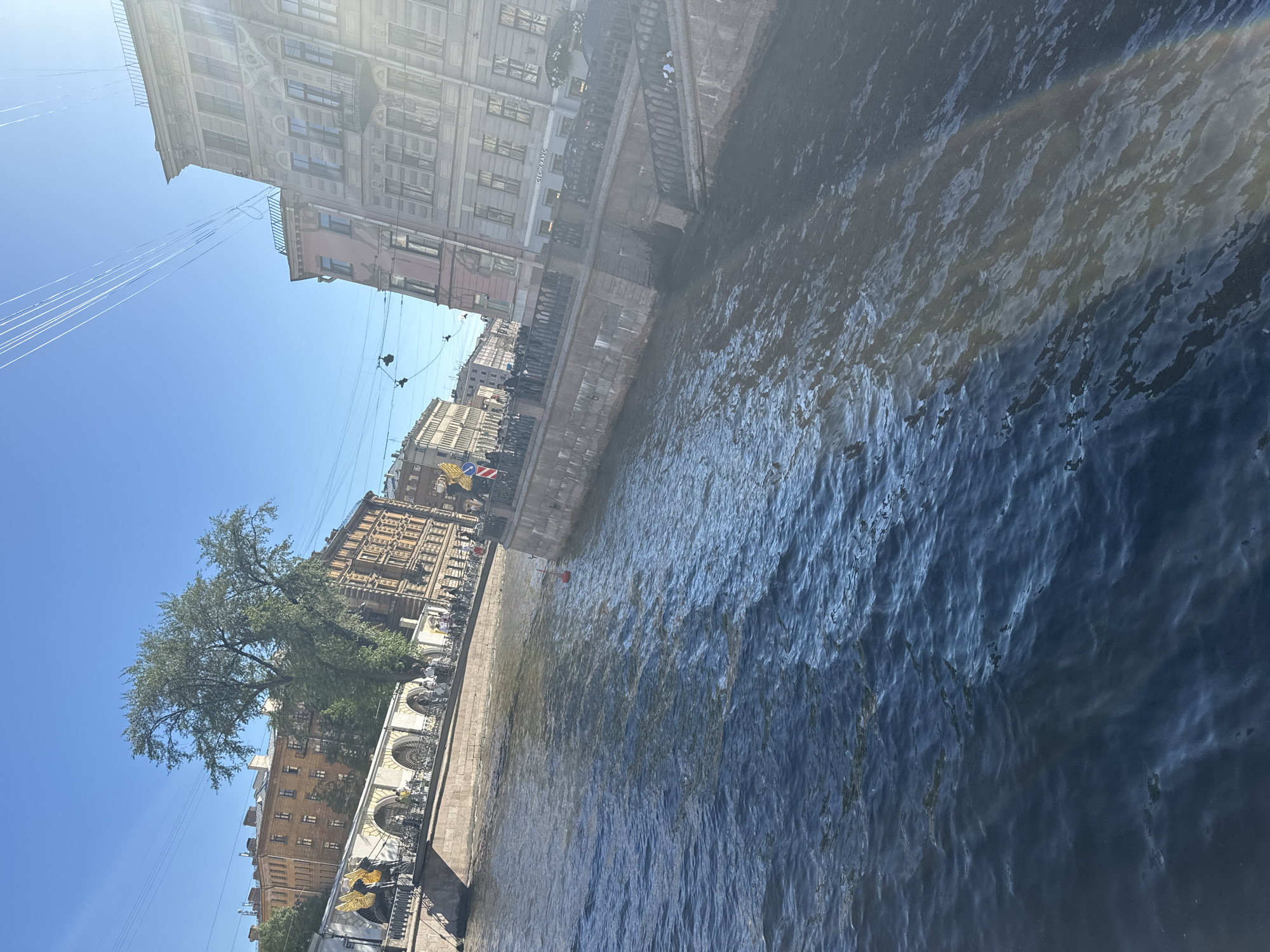 This is a photograph taken from a boat on a canal in Saint Petersburg, Russia, likely during the daytime under a clear blue sky. The image features buildings lining the canal's edge, with intricate architectural details and varying facades. One building appears to be a light pink color with multiple windows and balconies, while others are constructed from brick and stone. A large tree with green foliage is visible between the buildings. The canal water is dark and appears rippled, reflecting sunlight and creating streaks of light on its surface. The canal banks are lined with stone walls and ornate railings. Pedestrians are present along the canal walkways, some clustered together, suggesting public activity. There are also elements of infrastructure, such as overhead wires or cables, and a red buoy floating in the water. The perspective suggests the viewer is looking at an angle across the water towards the buildings and the canal's edge.
