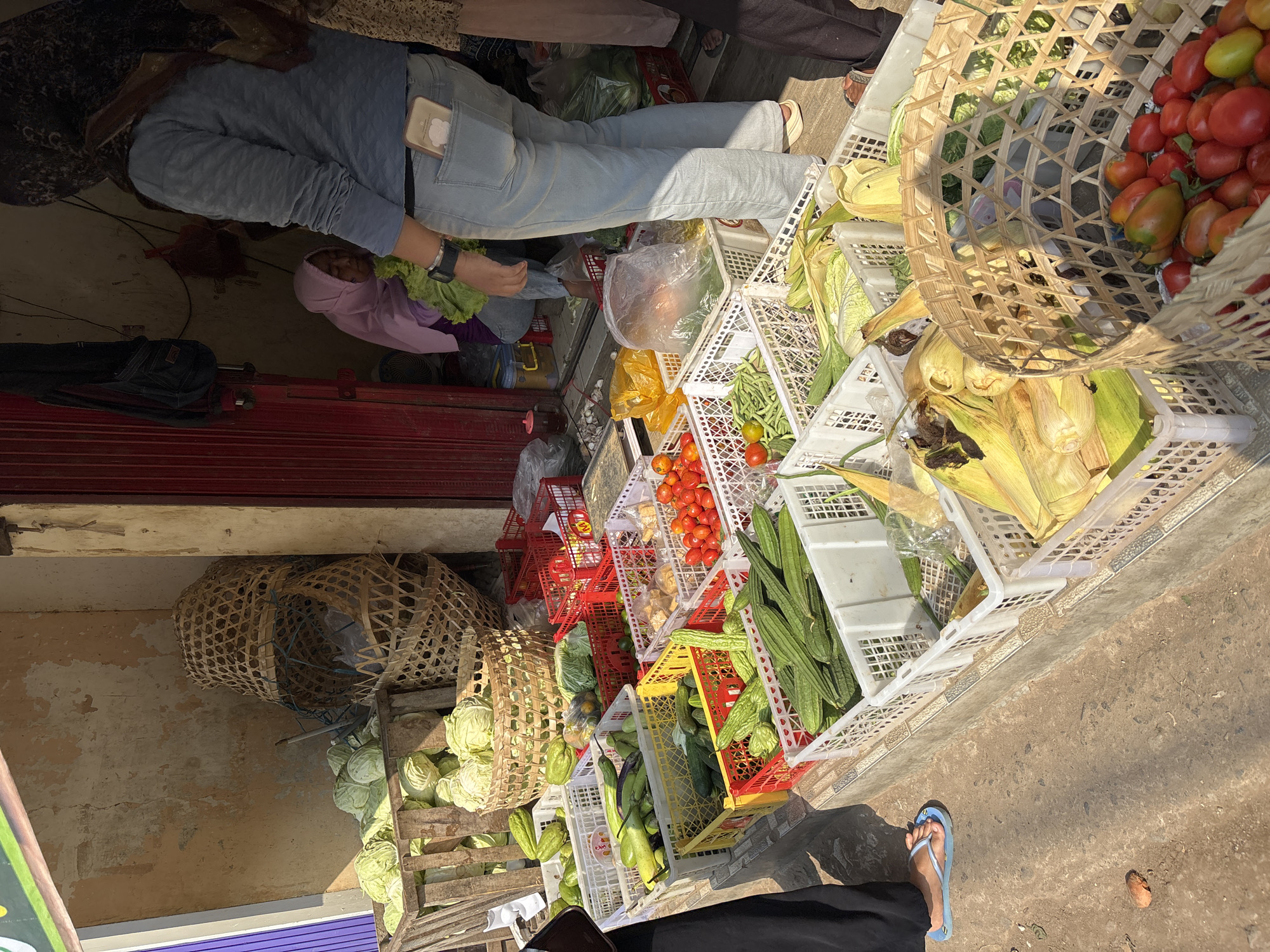 In Tirtomoyo, Indonesia, an outdoor market stall displays an abundance of fresh produce. Cabbages are stacked in a wooden crate to the left, with woven baskets of various sizes containing more vegetables and produce. White and red plastic crates are filled with items such as cucumbers, gourds, tomatoes, green beans, and corn. In the background, two women are partially visible, one wearing a purple headscarf and the other a patterned scarf. A woman in a blue long-sleeved shirt and jeans is standing in the foreground, with a smartphone visible in her back pocket. The ground is concrete, with a dirt area on the left side of the image. The market appears to be in full operation, with produce displayed for sale.