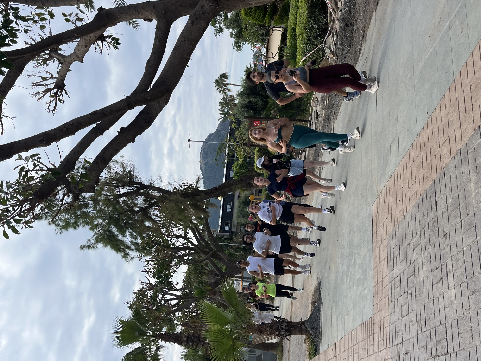 A group of approximately 13 people are engaged in an apparent exercise or running activity on a paved walkway. The group is staggered in a line, moving away from the camera's perspective. The setting appears to be a public park or a promenade in Alanya, Turkey.
The people are wearing a variety of athletic attire including shorts, t-shirts, tank tops and leggings. The clothing varies in color and some individuals are wearing athletic shoes. They seem to be jogging or running along the path.
The walkway is made of concrete slabs with a brick border. A large tree and other lush vegetation, including palm trees, frame the scene on the left. A mountain is visible in the background, near what appears to be a building. Above, the sky is partly cloudy.