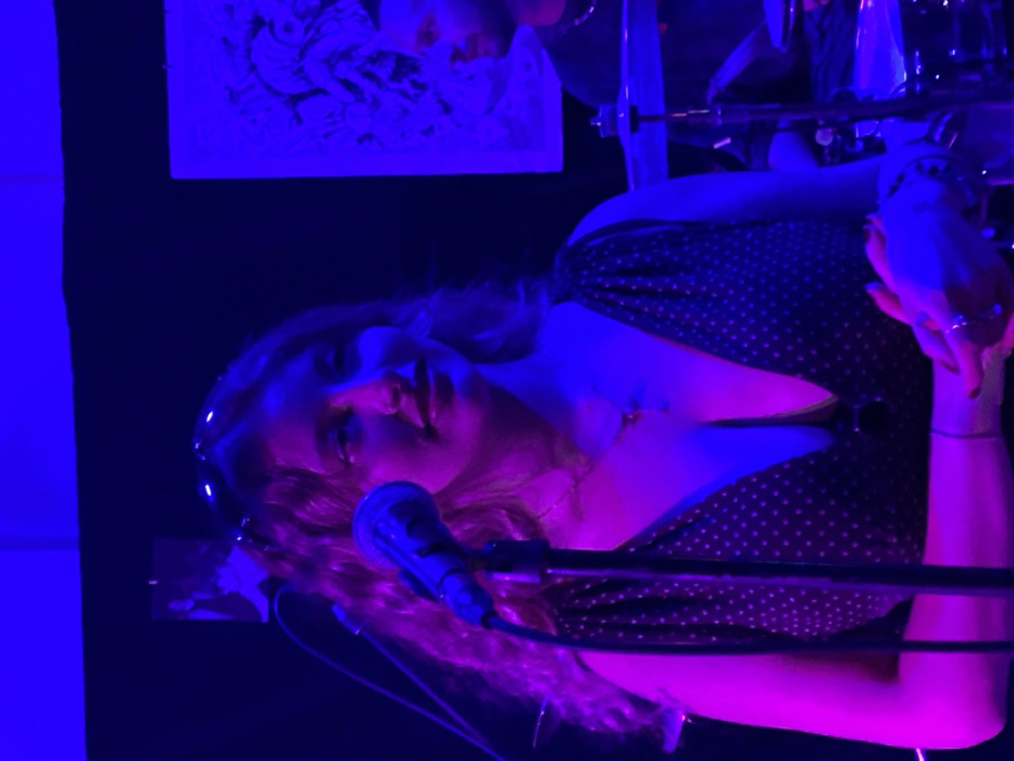The image captures an indoor performance scene illuminated by strong blue and magenta stage lighting. In the foreground, a young adult female with long, wavy light-colored hair stands slightly to the left of the center, her head turned left while her body faces slightly right. She wears sunglasses pushed up onto her head and a dark, polka-dotted dress with a deep V-neckline, adorned with a pendant necklace. Her hands are clasped in front of her lower torso, displaying rings and bracelets, and red nail polish on her left hand. A dark microphone on a stand is positioned directly in front of her. To the right and in the background, a young adult male is partially visible, seated behind a drum kit, including a visible cymbal and drum. He has dark hair, a short beard, wears a dark t-shirt, and a silver-colored chain necklace, and is looking towards the left, in the direction of the female subject. The background is a dark wall, featuring a small, dark-framed image of a person on the upper left and a larger, light-framed artwork with black line art on the upper right. The specific location within Buenos Aires, Argentina, is not identifiable from the visual information presented.