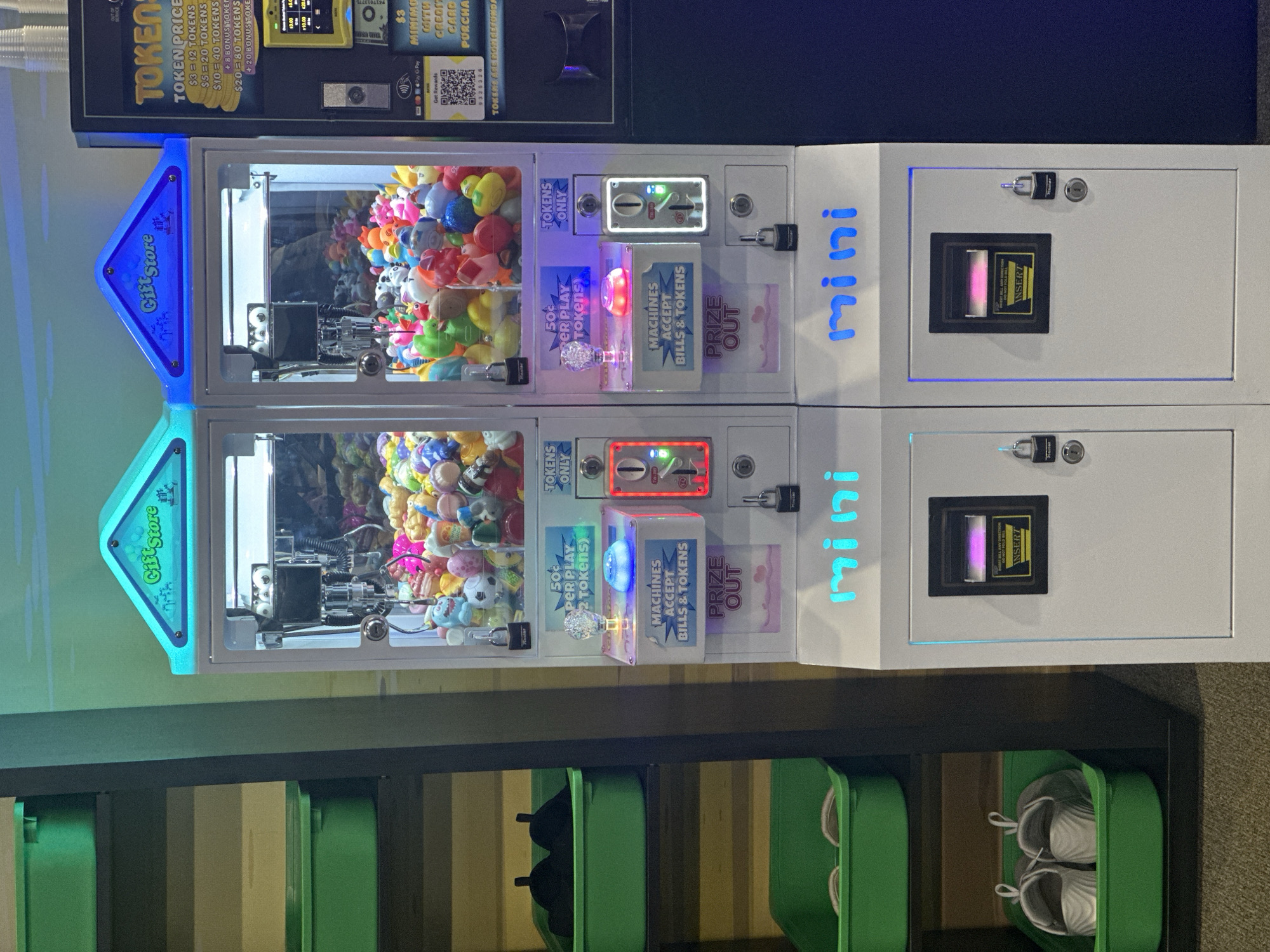 Two identical "Gift Store" brand mini crane machines, each with a blue neon sign on top, are positioned side-by-side in what appears to be an indoor arcade or entertainment venue. The machines are white with clear glass fronts showcasing an assortment of colorful plush toys. Each machine has coin slots, token slots, and a bill acceptor. Below the prize dispensing area, the word "mini" is illuminated in blue neon. To the left of the crane machines, shelves are filled with green plastic bins containing items such as shoes and what appear to be other retail goods. To the right of the crane machines is a dark blue wall. A token vending machine with a screen and various price options is visible to the far right. The overall environment is brightly lit. The location is specified as Buffalo Grove, United States of America.