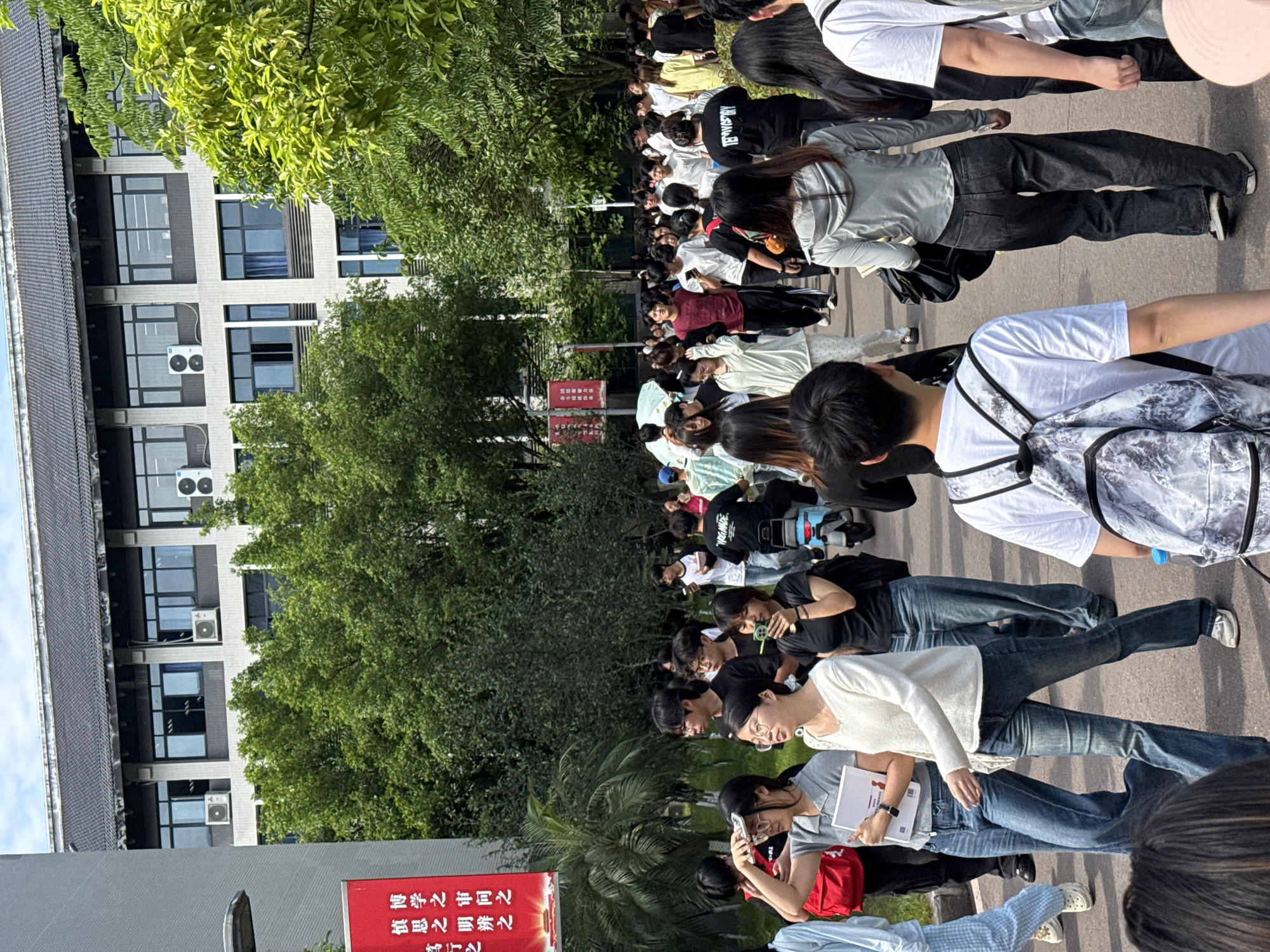 A crowd of people stands on a paved surface in front of a building with multiple windows and air conditioning units in Jiangjin, China. Trees flank the building. A red sign with Chinese characters is visible. The people are wearing a variety of clothing, including white shirts, black shirts, jeans, and jackets. Some are holding phones or notebooks.