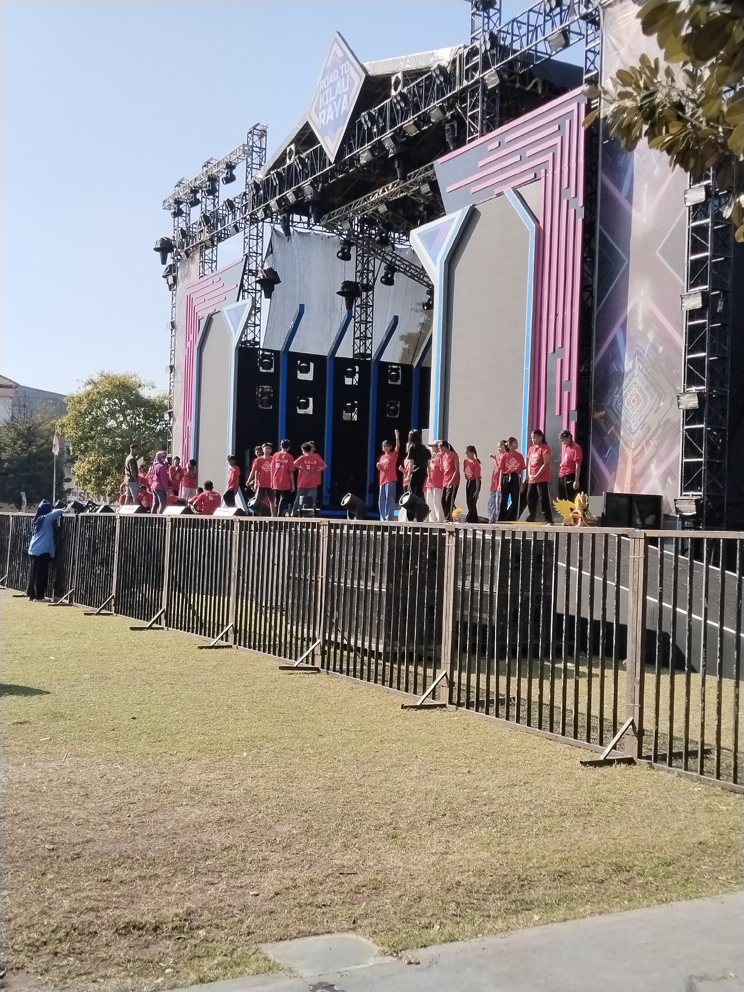 This image captures an outdoor event with a large stage set up for a performance. The stage features massive LED screens and intricate scaffolding, suggesting a significant production. A group of individuals, mostly young people dressed in red t-shirts and black pants, are gathered on the stage. Some are standing, while others are seated or in motion, possibly rehearsing or waiting for their turn. In front of the stage, a black metal fence encloses the performance area, with a grassy lawn beyond it. The overall scene suggests a public gathering or festival. While the location is identified as Kecamatan Pati, Indonesia, specific landmarks or environmental details that would confirm this location are not readily apparent in the image itself. The sky is clear and blue, indicating fair weather.
