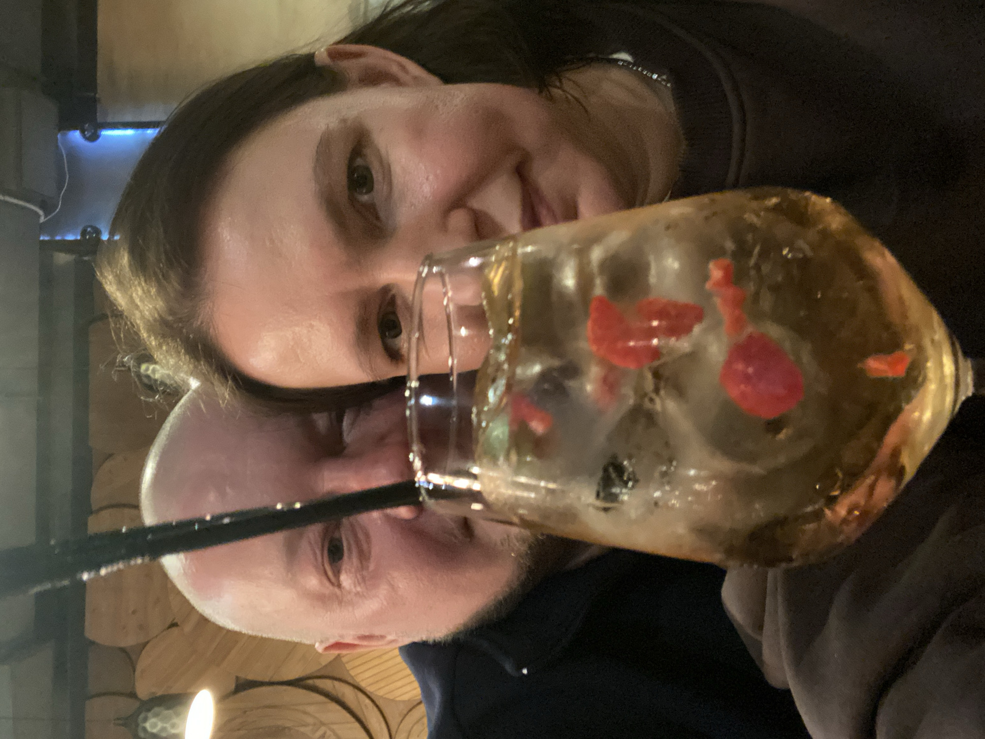 A selfie depicts two individuals posing with a cocktail glass. The glass contains ice, a clear liquid, and red fruits. A black straw protrudes from the glass. The person closest to the camera on the right has brown hair and is wearing a dark-colored top. The other person, slightly behind, is bald and wearing a dark blue top. The background includes a wall with a wood paneling effect, and a dimly lit fixture. The location is possibly in Yekaterinburg, Russia.