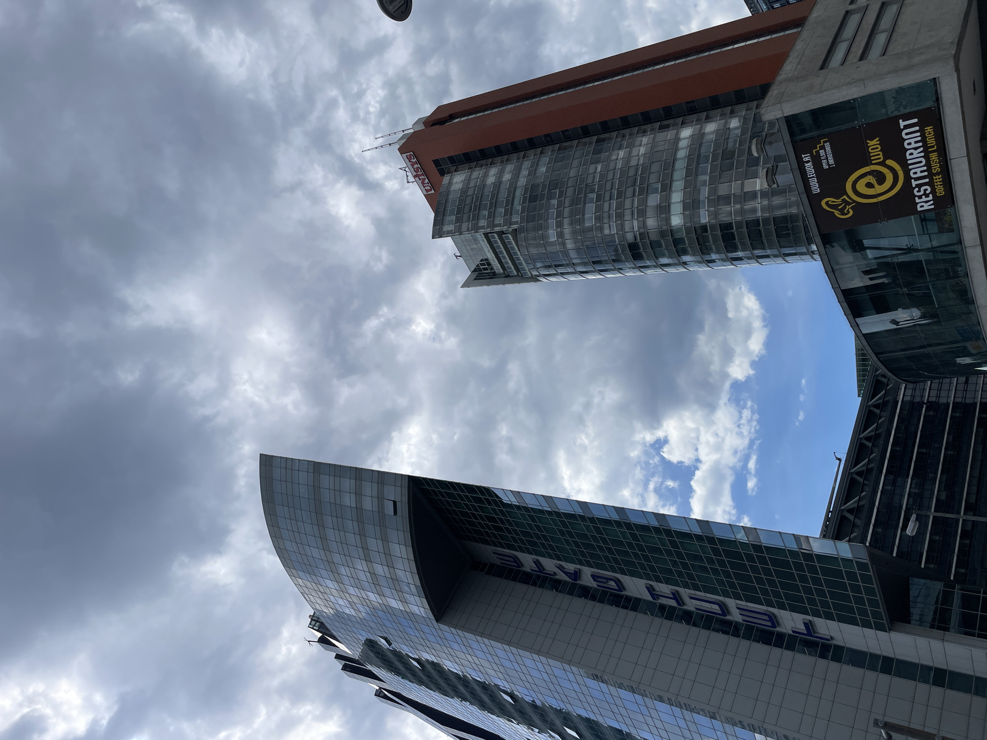The image shows a low-angle view of two buildings in an urban setting, likely in Vienna, Austria, due to the presence of signage in German. The building on the right, the Unisys building, has a reddish-brown upper section and a curved glass facade on the lower part. Adjacent to it is a sign for "e-Wok Restaurant" and "Coffee Sushi Lunch" with the website "www.ewok.at" printed on the sign. The building on the left has a modern, curved design with the words "TECH GATE" visible in large lettering. The sky is overcast with a mix of dark and light clouds. There is a streetlight in the upper-left corner.
