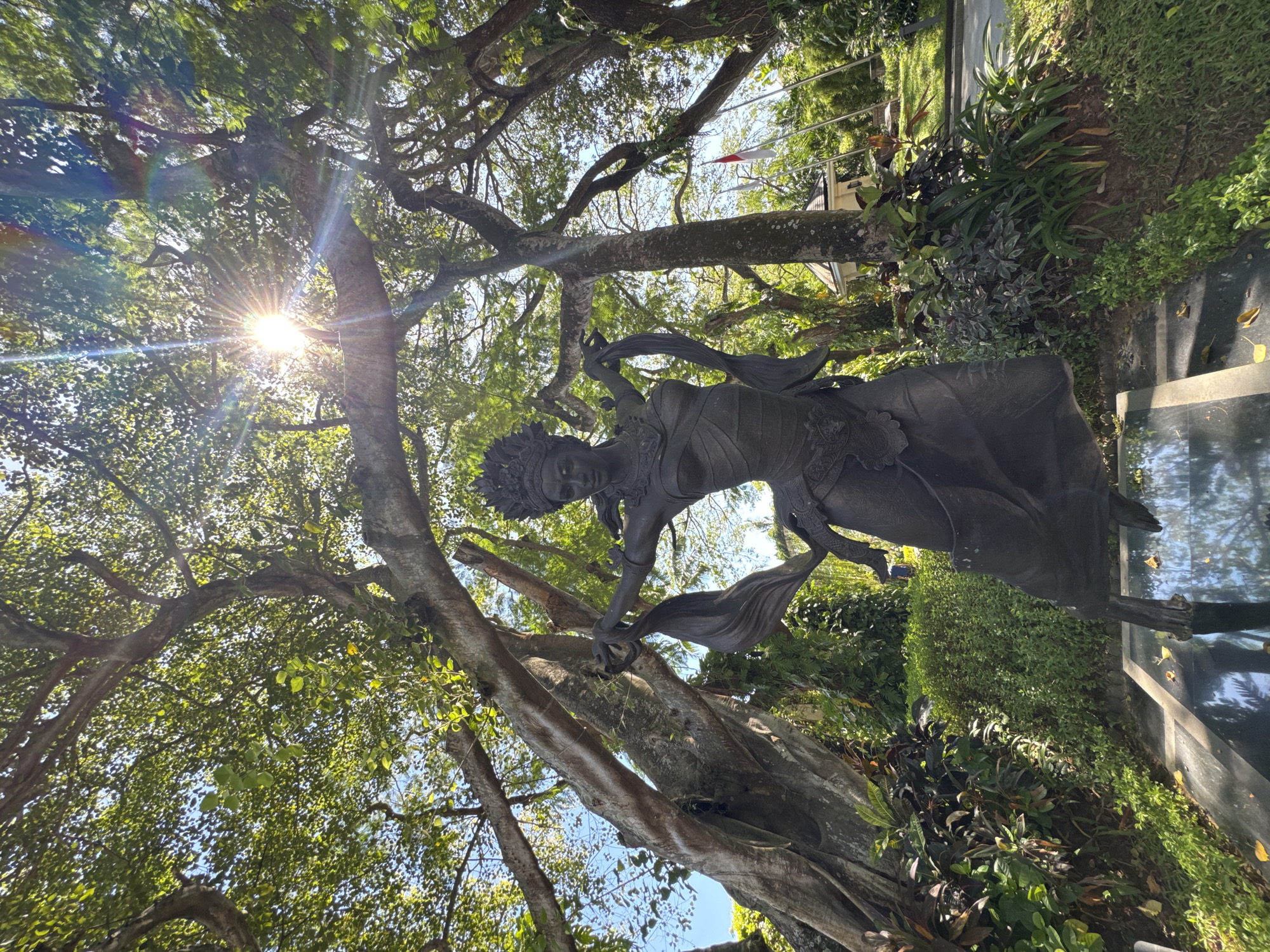 The image depicts a statue in Kuta Selatan, Indonesia. The statue appears to be bronze or a dark metal, depicting a woman in traditional dress with flowing fabric. The statue is positioned amongst a large tree with thick branches and green foliage, with sunlight filtering through the leaves. Other greenery and plant life surround the base of the statue. A paved area and possibly a building are visible in the background. The Indonesian flag is flying in the distance.