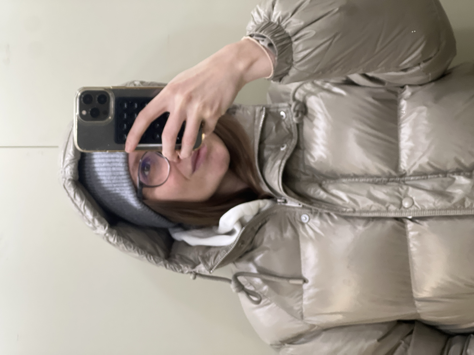 An upright mirror selfie captures a person taking a photo with a smartphone. The subject, likely female, wears a light grey knitted beanie and round, wire-rimmed glasses. Her medium-brown hair is visible beneath the hat. She holds a dark grey smartphone, likely an iPhone with a triple-lens camera system visible through a clear case, in her hand, which also displays a thin bracelet on the wrist.
She is dressed in a shiny, light taupe or beige puffer jacket with a hood, featuring horizontal quilting. A white layer, possibly a hoodie or undershirt, is visible beneath the jacket collar. Her lips are slightly parted in a neutral expression. The background consists of a plain, light-colored wall, indicating an indoor setting. The choice of warm clothing suggests cool or cold weather, consistent with the provided location context of Khimki, Russia, although no specific location cues are visible in the image itself. No text is visible in the image.
