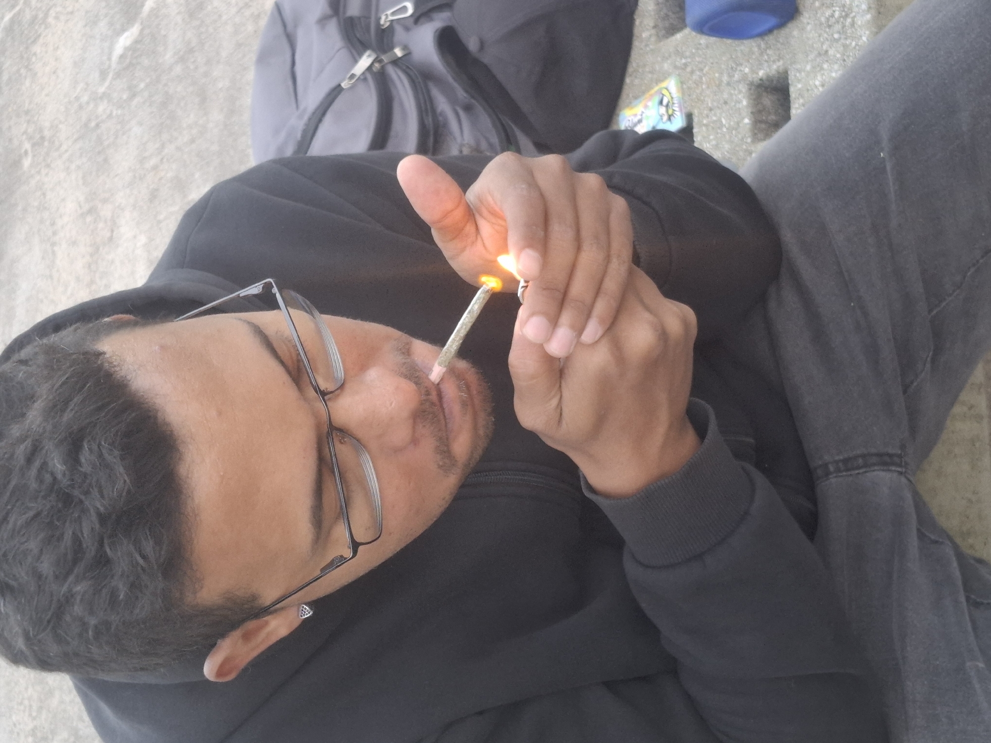A person with dark hair, glasses, and a diamond earring is lighting a marijuana cigarette with a lighter. The person is wearing a dark-colored hoodie and gray jeans. A gray backpack and a blue can are visible in the background. The scene appears to be outdoors on a concrete surface. The specific location in Buenos Aires, Argentina is indiscernible.