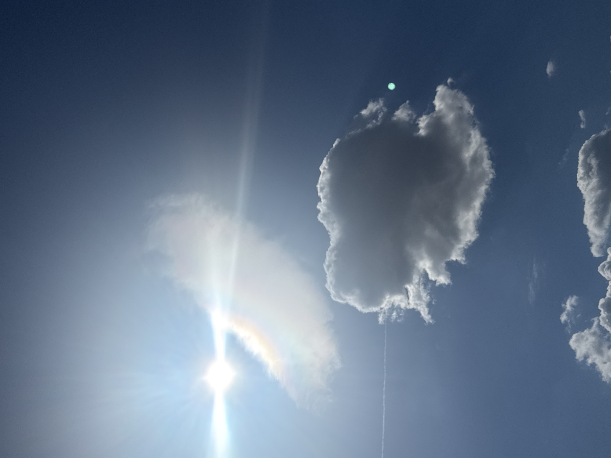 The image shows a daytime sky with several clouds. A bright sun is visible on the left, creating a lens flare effect. A cloud partially obscures the sun and displays a faint rainbow effect. Another cumulus cloud is in the upper center of the frame. Another cloud is to the far right of the frame. A contrail from a jet is below the cumulus cloud in the center of the frame. The sky is blue. The location is in Lupanovo, Russia.