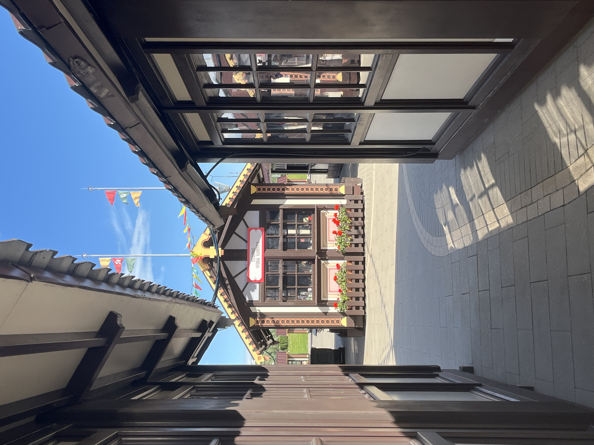 This is a photograph of a pedestrian walkway between two dark brown wooden buildings. The buildings have awnings with dark brown trim and tiled roofs. In the distance, a small, quaint building with a peaked roof and decorative trim is visible, flanked by colorful triangular flags. The building has windows with brown frames and a red sign with white text. Red flowers are planted in wooden planters in front of this building. The walkway is paved with gray rectangular tiles and is illuminated by sunlight, casting long shadows from the buildings. A clear blue sky with a few scattered clouds is visible above. The scene is clean and well-maintained. The architecture suggests a fairground, market, or a themed area within a larger establishment. The provided metadata indicates the location is in Nizhny Novgorod, Russia. No people are present in the image.