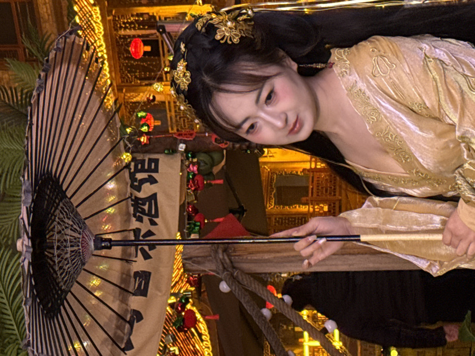 An East Asian female subject is depicted at night in an illuminated urban setting, consistent with a tourist area such as Hongyadong in Yuzhong District, China. The subject, positioned centrally and facing to her right, wears a gold-colored, V-neck traditional Chinese-style gown with embroidered detailing. Her dark hair is styled with gold-toned floral and pearl headdresses and she wears matching dangle earrings. Her makeup includes pronounced pink blush on her cheeks and eyelids. She holds a black and translucent beige traditional paper parasol with her right hand.
Behind the subject, a beige banner with dark Chinese characters "自木酒馆" (Zì Mù Jiǔ Guǎn) is visible above the parasol. The background features multi-story traditional wooden architecture adorned with numerous warm-toned string lights and red Chinese lanterns. To the left, a thick braided rope, strung with small white lights, acts as a railing. A blurred male figure in a dark hooded jacket is partially visible in the left background. The scene is brightly lit, indicating a festive nighttime environment.