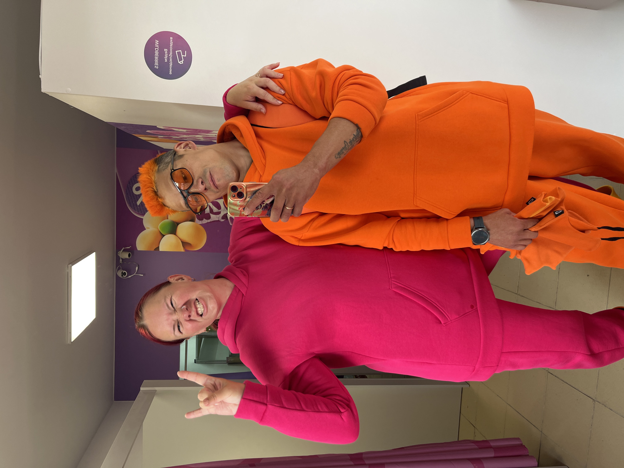 Two individuals are posing for a selfie in what appears to be a clothing store fitting room.
A person with orange hair and eyeglasses is wearing an orange hoodie and pants. The person is holding a smartphone, likely taking the photo, and has a tattoo on their arm and a wristwatch on their wrist. The second person is wearing a pink hoodie and pants and is making a hand gesture with two fingers extended.
The fitting room has purple wallpaper with fruit images, and there is a white sign with Cyrillic writing. A light fixture is visible on the wall. The scene is in a store location in Chaikovsky, Russia.