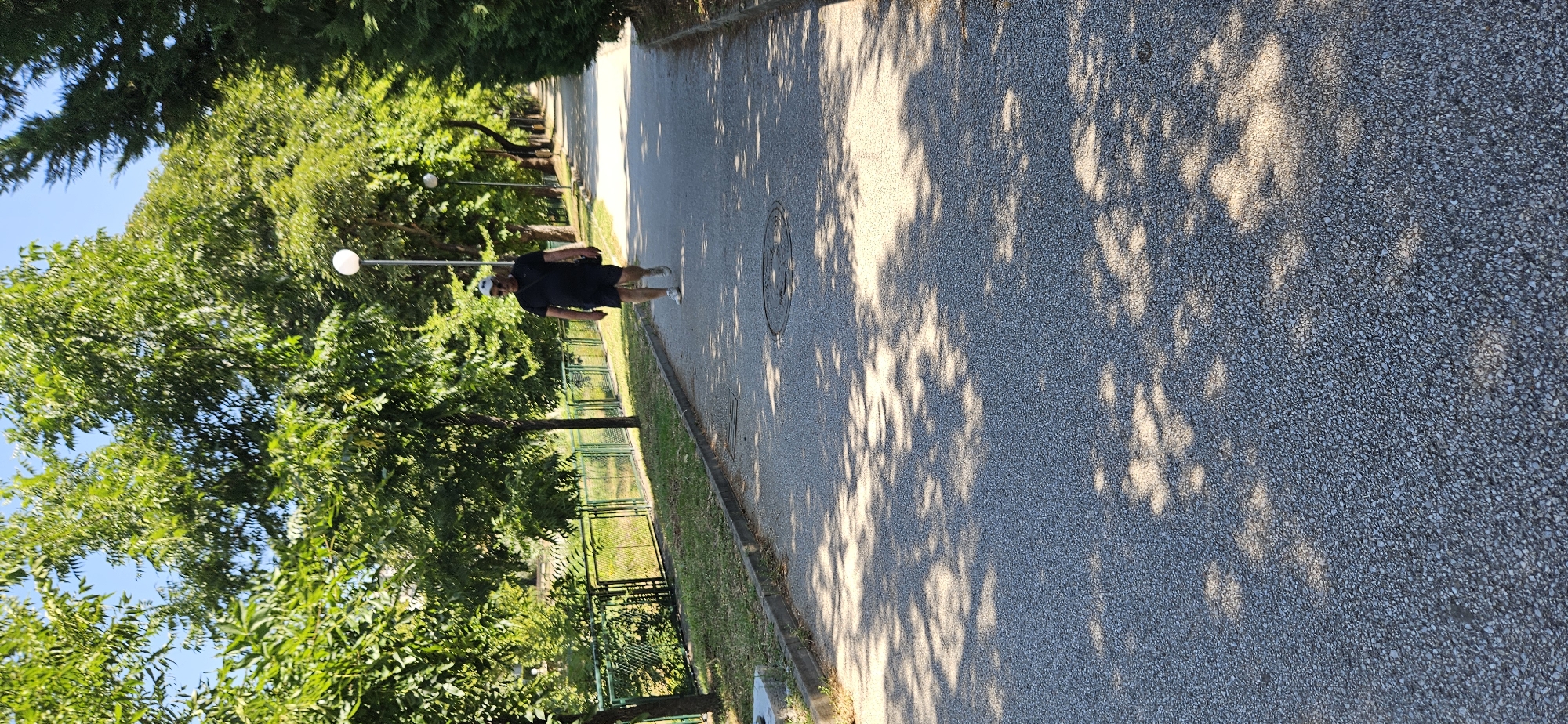 A man in a black t-shirt, black shorts, a white baseball cap, sunglasses, and white sneakers walks on a paved pathway. He carries a dark bag slung over his left shoulder. The pathway is lined with trees and grass, with a metal fence bordering a grassy area to the left. A light post stands near the man. The pathway is dappled with sunlight and shadows. The scene is set on a paved pathway in a park-like setting, likely within the city of Blagoevgrad, Bulgaria, given the context.