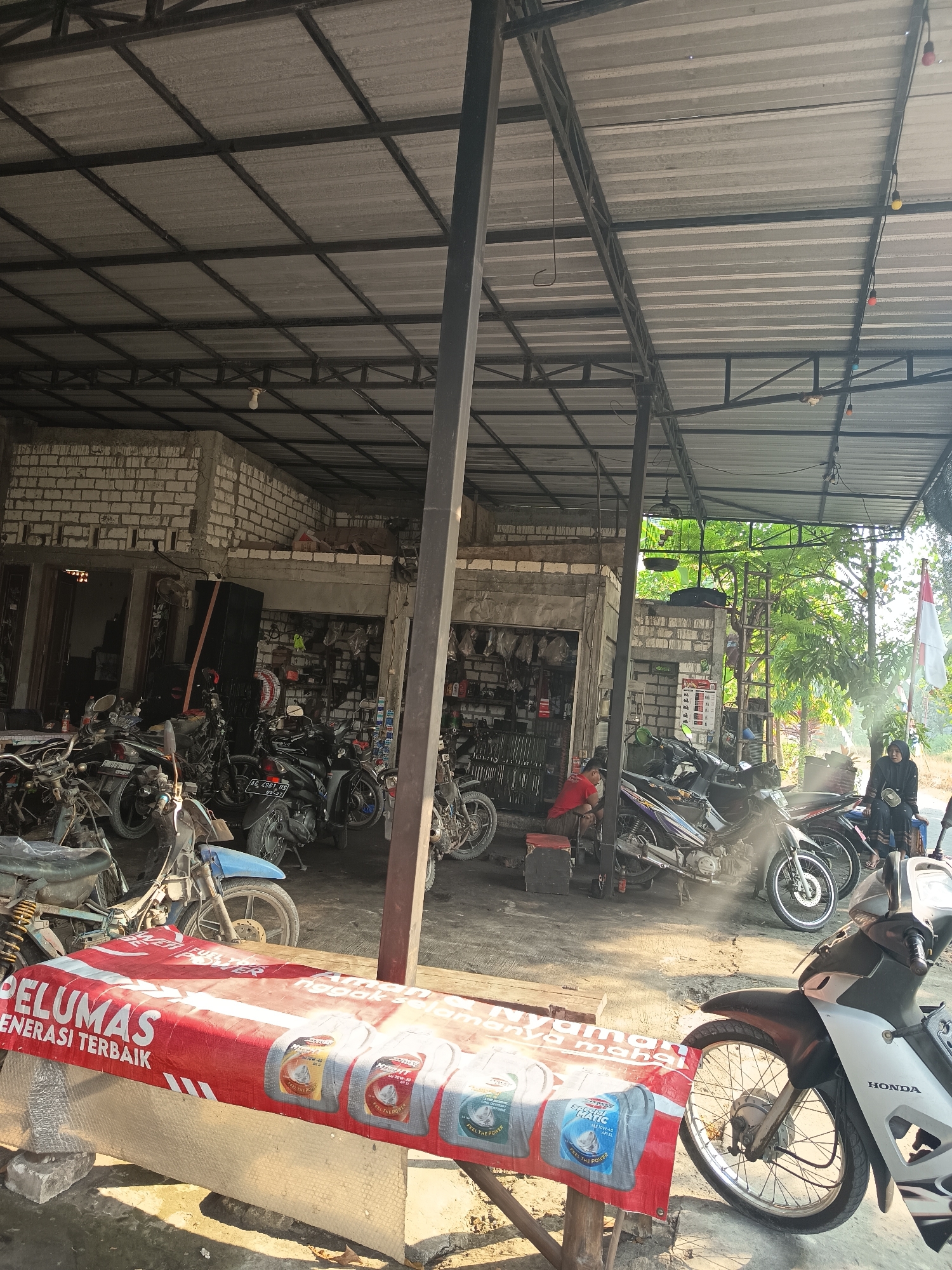 The image depicts an outdoor motorcycle repair shop in Sugio, Indonesia. The scene is dominated by a metal-framed structure with a corrugated metal roof, providing shade over several motorcycles. In the foreground, a red banner with white text, possibly an advertisement, lies across a makeshift table. Several motorcycles, some appearing to be under repair or for sale, are parked around the shop. The background shows a building constructed with white bricks, partially open, revealing shelves stocked with motorcycle parts. A few individuals are visible: one person in a red shirt is seated near some motorcycles, and another person in black clothing is sitting on a stool near the shop's entrance. A flag, likely the Indonesian flag, is flying on a pole to the right. The ground is a dirt and concrete mix, typical of an outdoor workshop environment. The lighting suggests it is daytime, with sunlight casting shadows and creating a hazy atmosphere, possibly due to dust or heat.