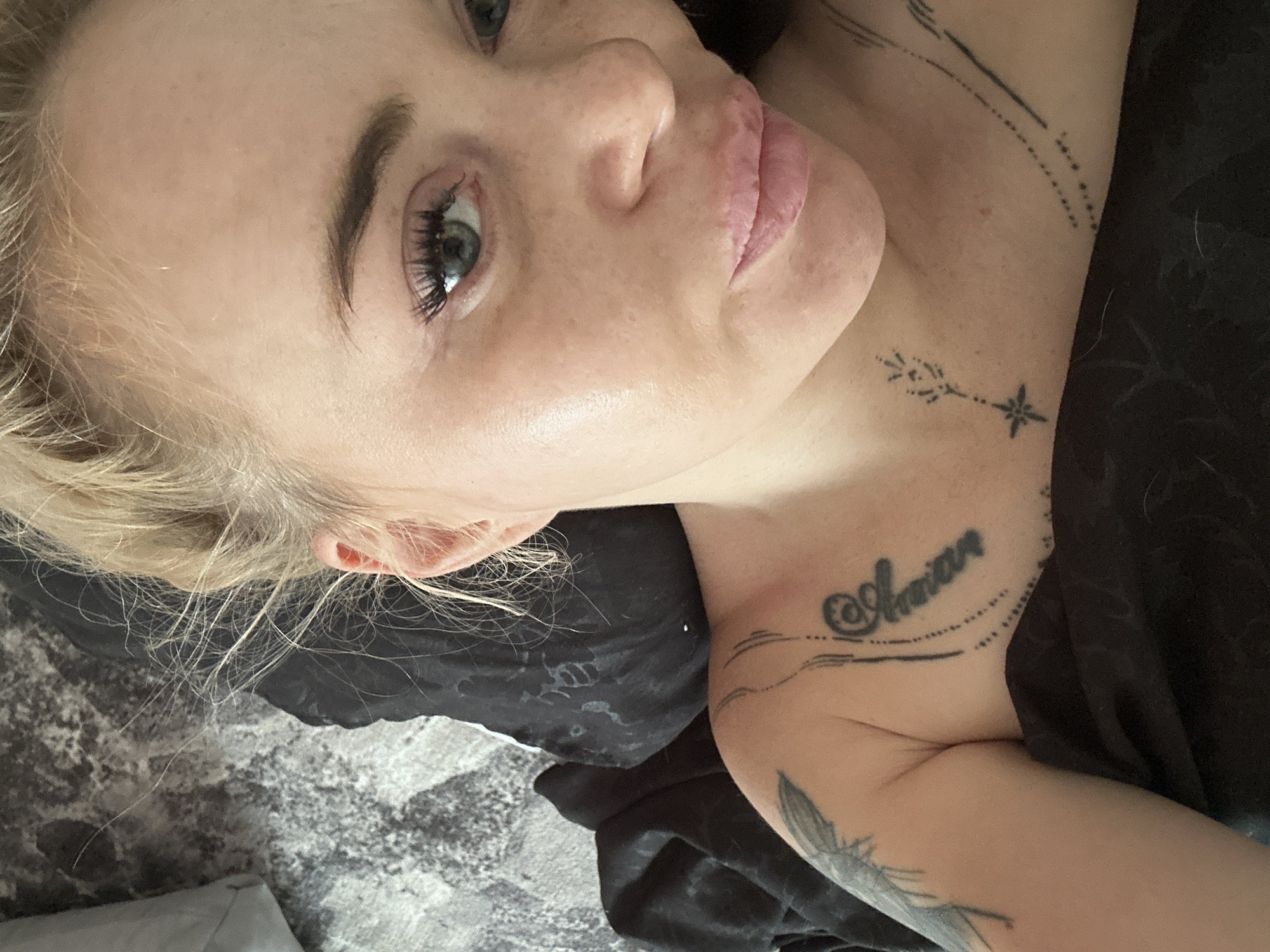 A fair-skinned individual with blonde hair tied in an updo, exhibiting visible flyaways, is depicted in a close-up, high-angle selfie. The subject's face is oriented towards the camera, featuring light-colored eyes, defined eyebrows, and enhanced eyelashes. Subtle skin texture and a slight sheen are present on the cheeks. The lips are full and light pink. The individual's upper torso is partially visible, covered by dark, solid-colored fabric, likely a blanket or sheet. Several tattoos are present: a faint winged or feather design on the left upper arm, a script-like text reading "Amnian" (approximate) encircled by dots on the right chest, a stylized arrow/plant design with a star at its base on the right collarbone, and a linear dotted pattern extending from the left shoulder area. The background consists of a dark grey and white mottled or abstract patterned surface, with a lighter grey object partially visible in the upper left. The scene is set indoors, in the city of Unknown, Unknown.