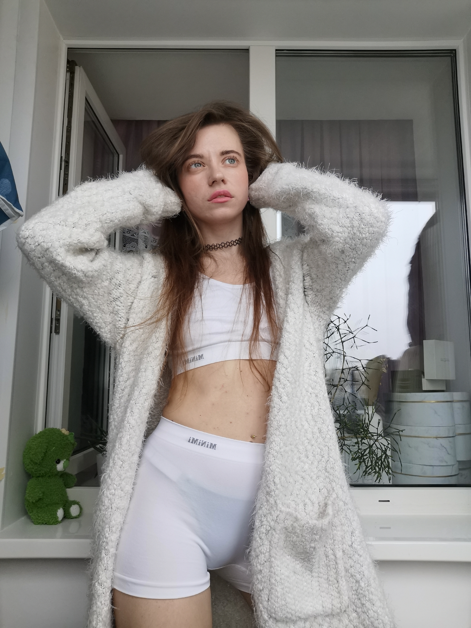 A young woman stands in front of a large window, posing for a photo. She wears a white sports bra, matching high-waisted shorts, and a long, fuzzy white cardigan. Her brown hair is slightly tousled, and she has a choker around her neck. Her hands are raised, seemingly adjusting her hair.
To the left of the woman, a small green stuffed animal sits on the windowsill. Behind her, the window reflects an outdoor scene, possibly a cityscape or a cloudy sky. A potted plant and several decorative boxes are also visible on the windowsill behind her. The location appears to be an indoor setting with natural light coming through the window. The location is Unknown, Unknown.