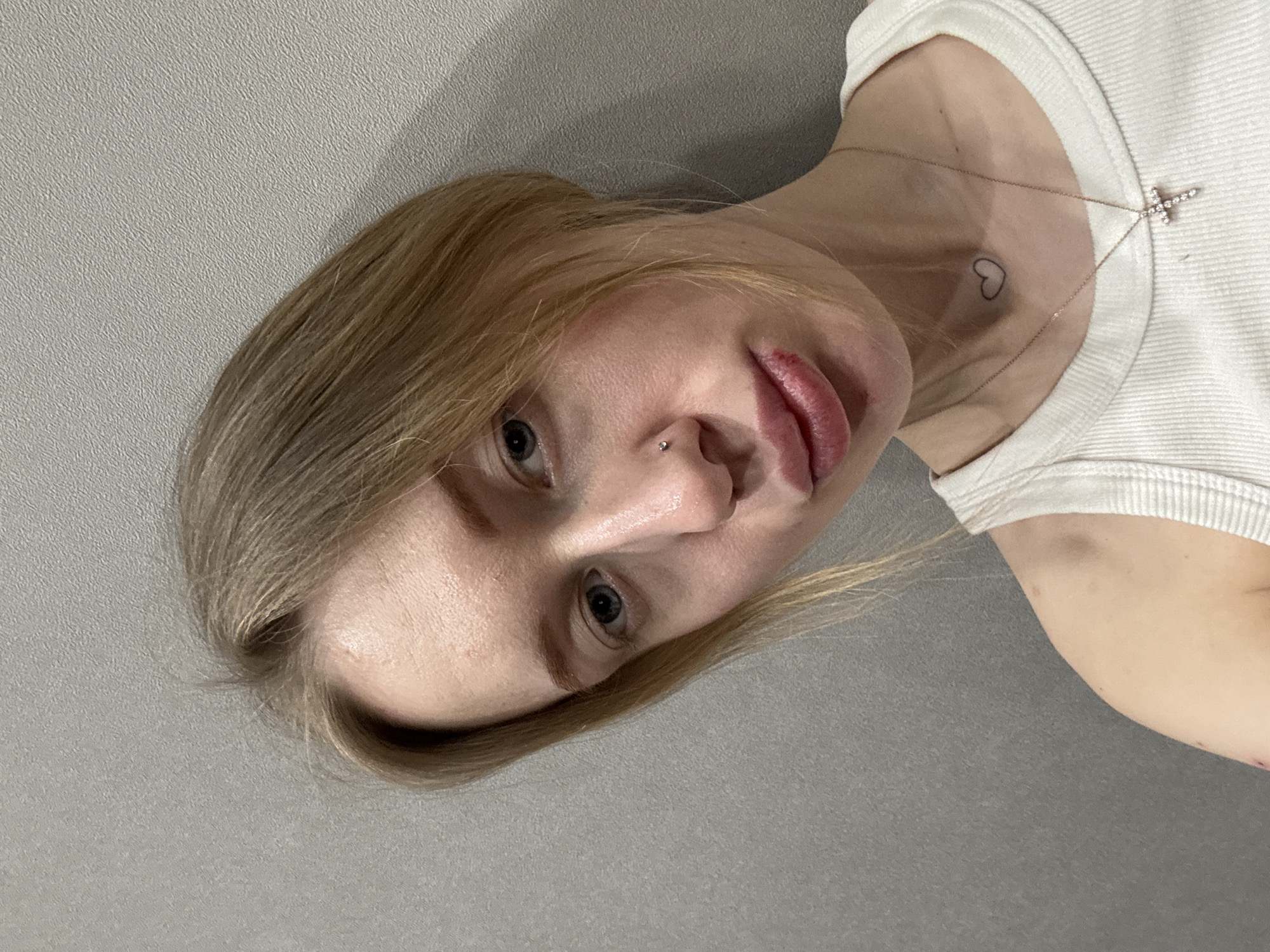 An indoor, chest-up portrait depicts a young adult female with blonde hair and light-colored eyes, positioned against a light grey, textured wall in what is reported as Rostov-on-Don, Russia. She is wearing a white ribbed tank top. A silver stud is present in her left nostril. Around her neck, a thin gold-colored chain supports a small cross pendant adorned with clear stones. A small outline heart tattoo is visible on her lower neck. Her lower lip displays a distinct dark reddish-brown lesion or scab. Additionally, a reddish abrasion or mark is visible on her left shoulder. The subject's gaze is directed towards the camera, and her expression is neutral. The scene is illuminated by soft, even lighting.