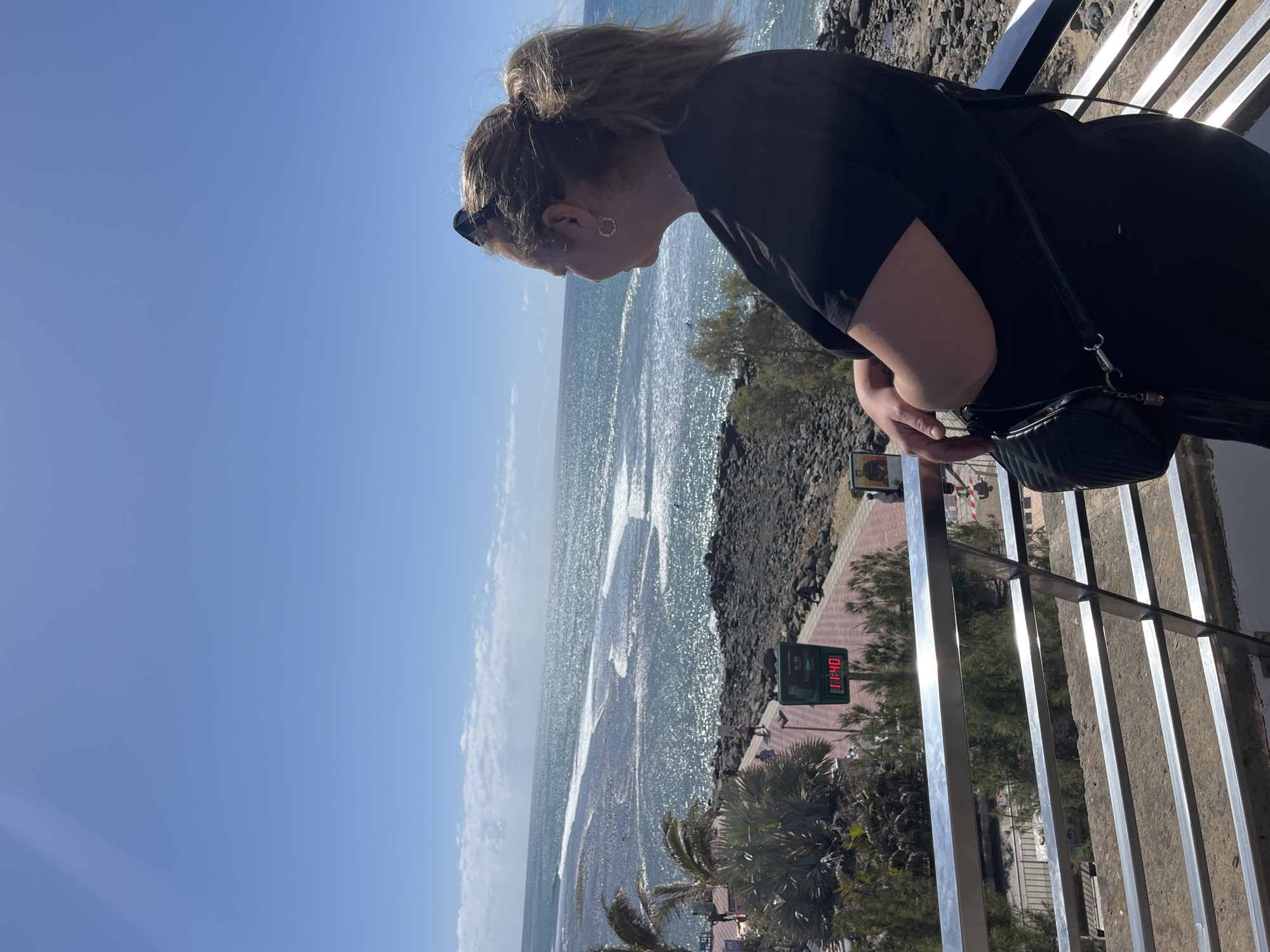 A woman with blonde hair pulled back in a bun stands by a silver railing, gazing out at the ocean in Maspalomas, Spain. She wears a black shirt and has a black purse hanging from her shoulder. The ocean sparkles with sunlight, and the sky is a clear blue. Below, a stone beach is visible next to a paved walkway with palm trees and a digital sign displaying the time.